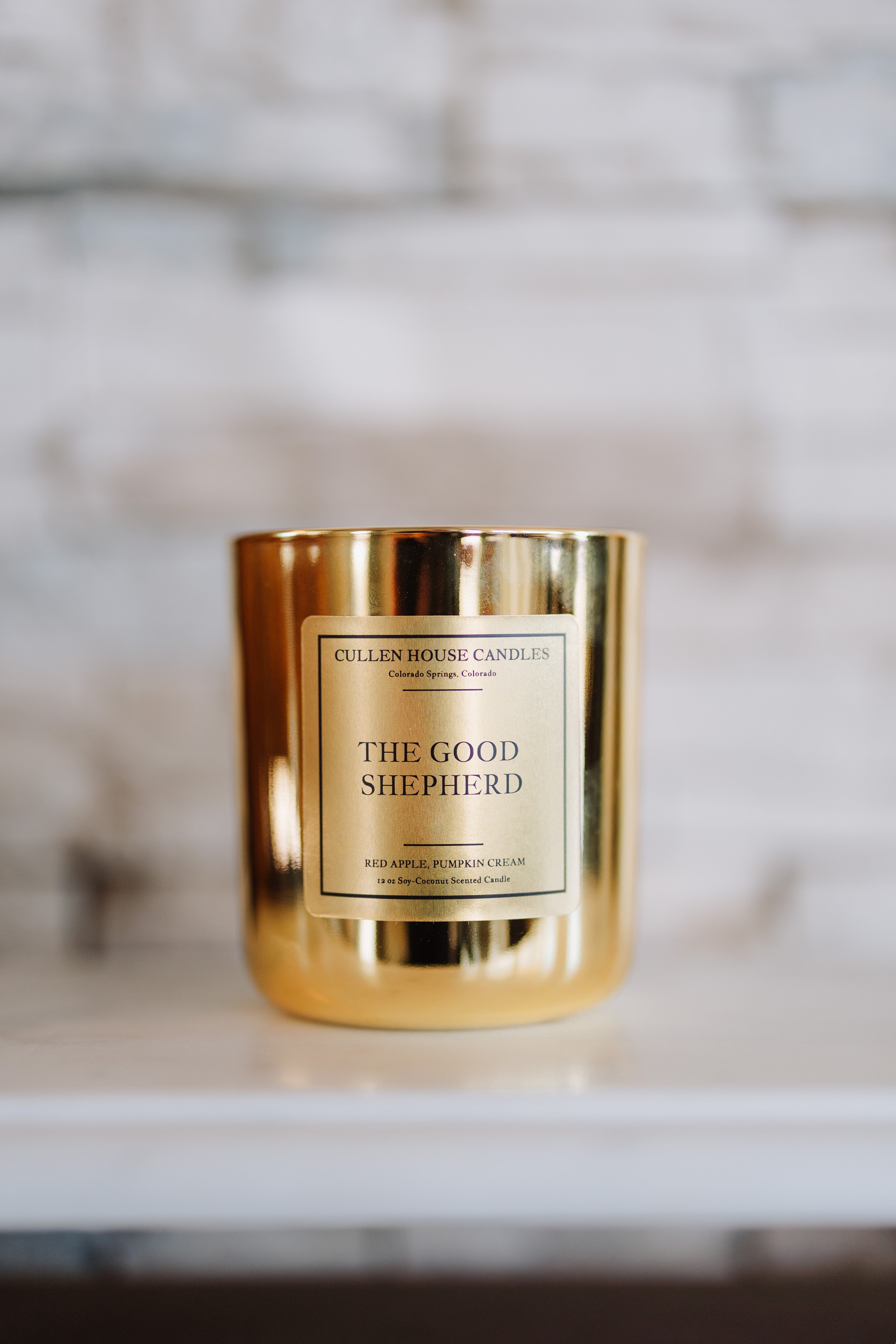 Gold candle jar labeled 'The Good Shepherd' by Cullen House Candles on a marble surface.