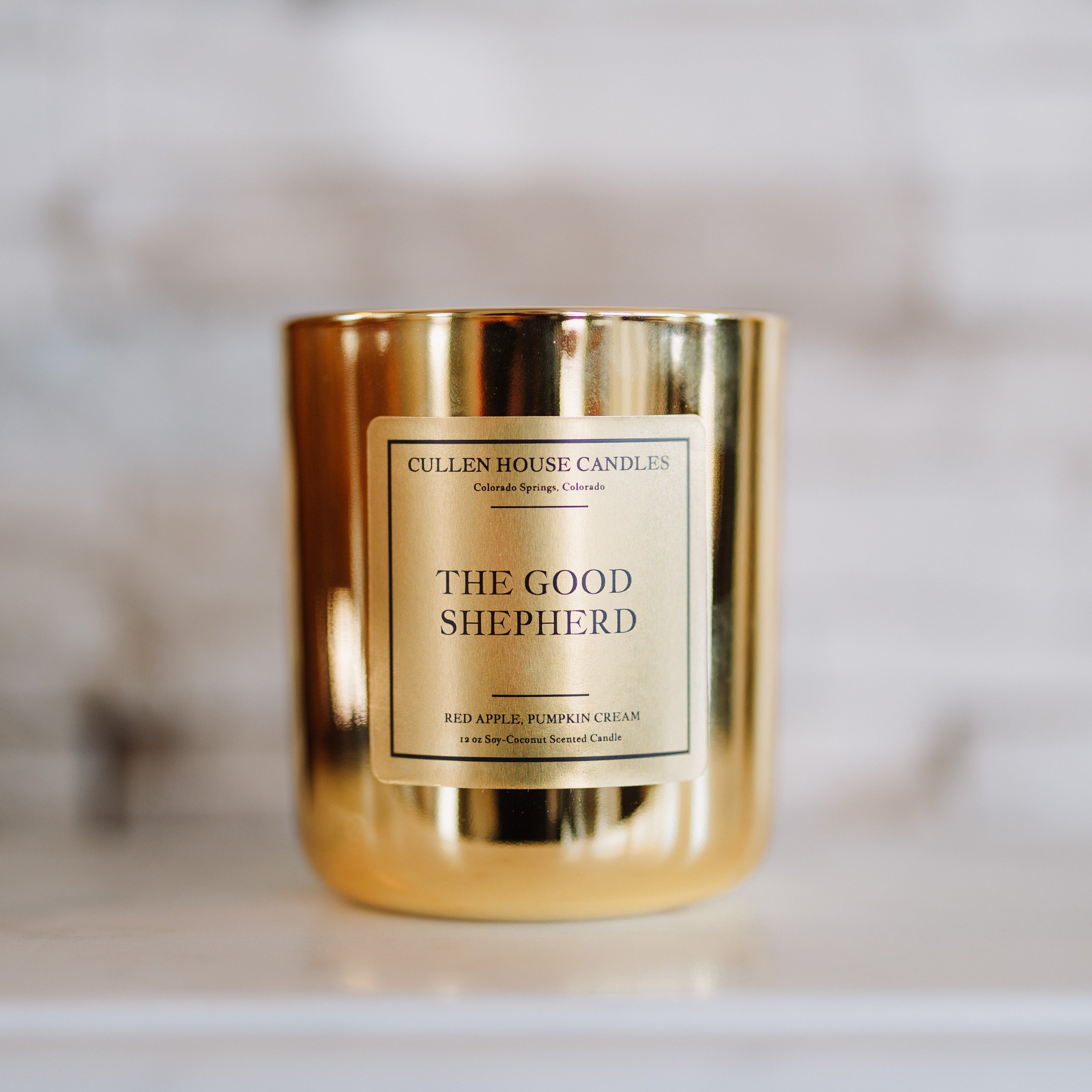 Gold candle jar labeled 'The Good Shepherd' by Cullen House Candles on a marble surface.
