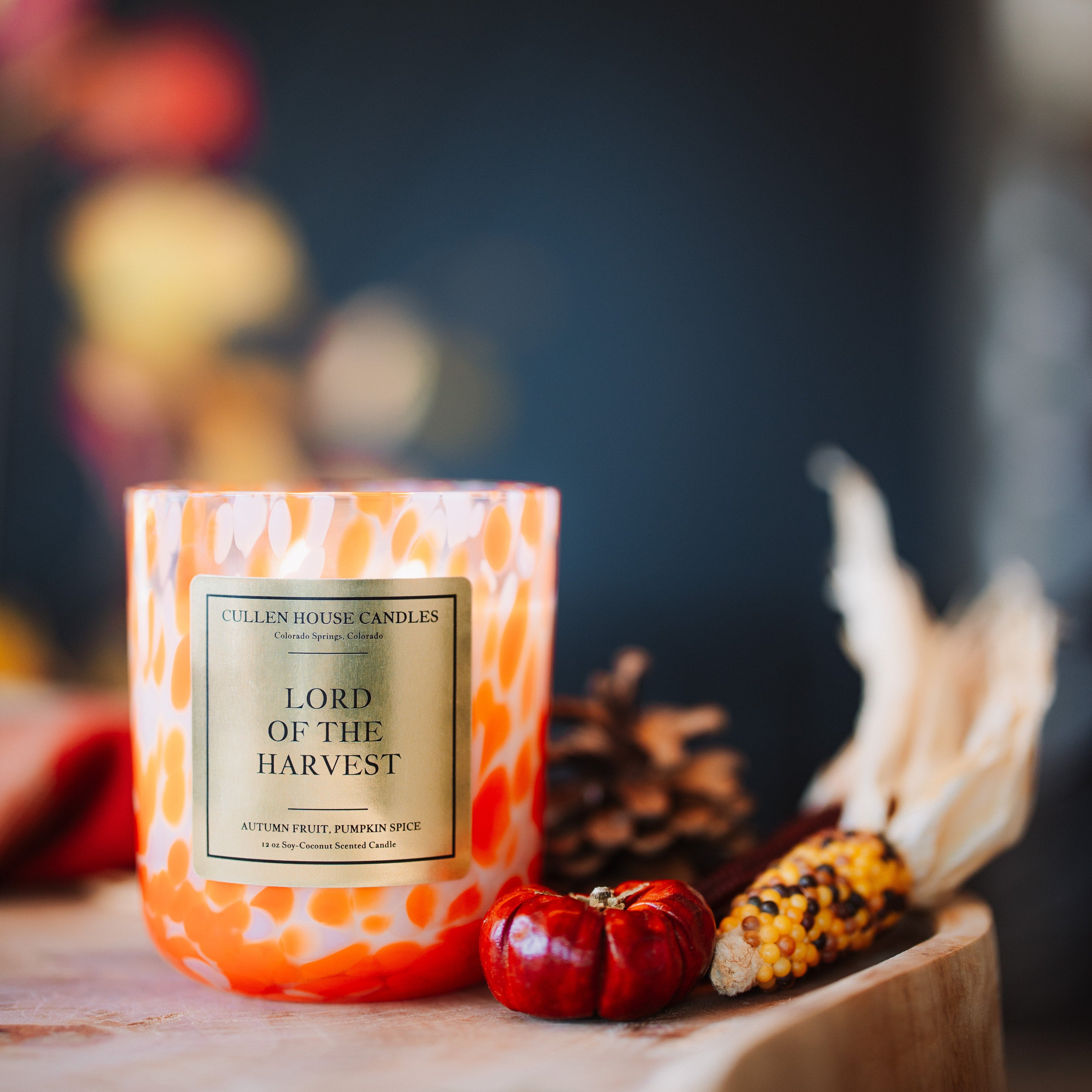 A jar candle labeled 'Lord of the Harvest' by Candle Shop Candles, styled on a wooden surface with autumn props including a small red pumpkin, dried corn, and leaves against a dark blurred background.