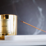 Gold candle labeled 'Prayer' with a smoking incense stick on a dark background