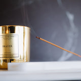 Gold candle labeled 'Prayer' with a smoking incense stick on a dark background