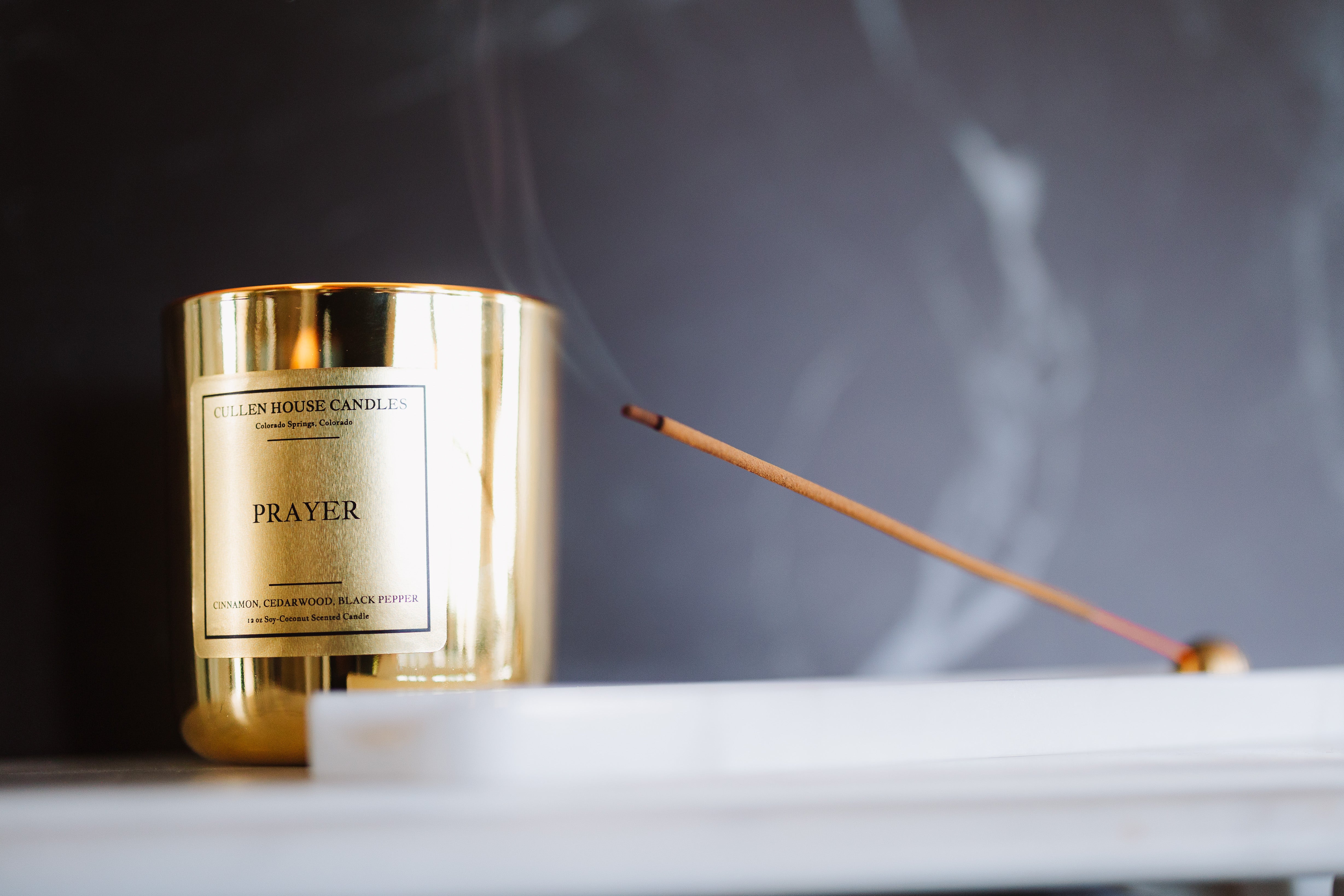 Gold candle labeled 'Prayer' with a smoking incense stick on a dark background