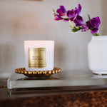 Candle with a gold label on a decorative stand next to a vase with purple flowers.