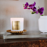 Candle with a gold label on a decorative stand next to a vase with purple flowers.