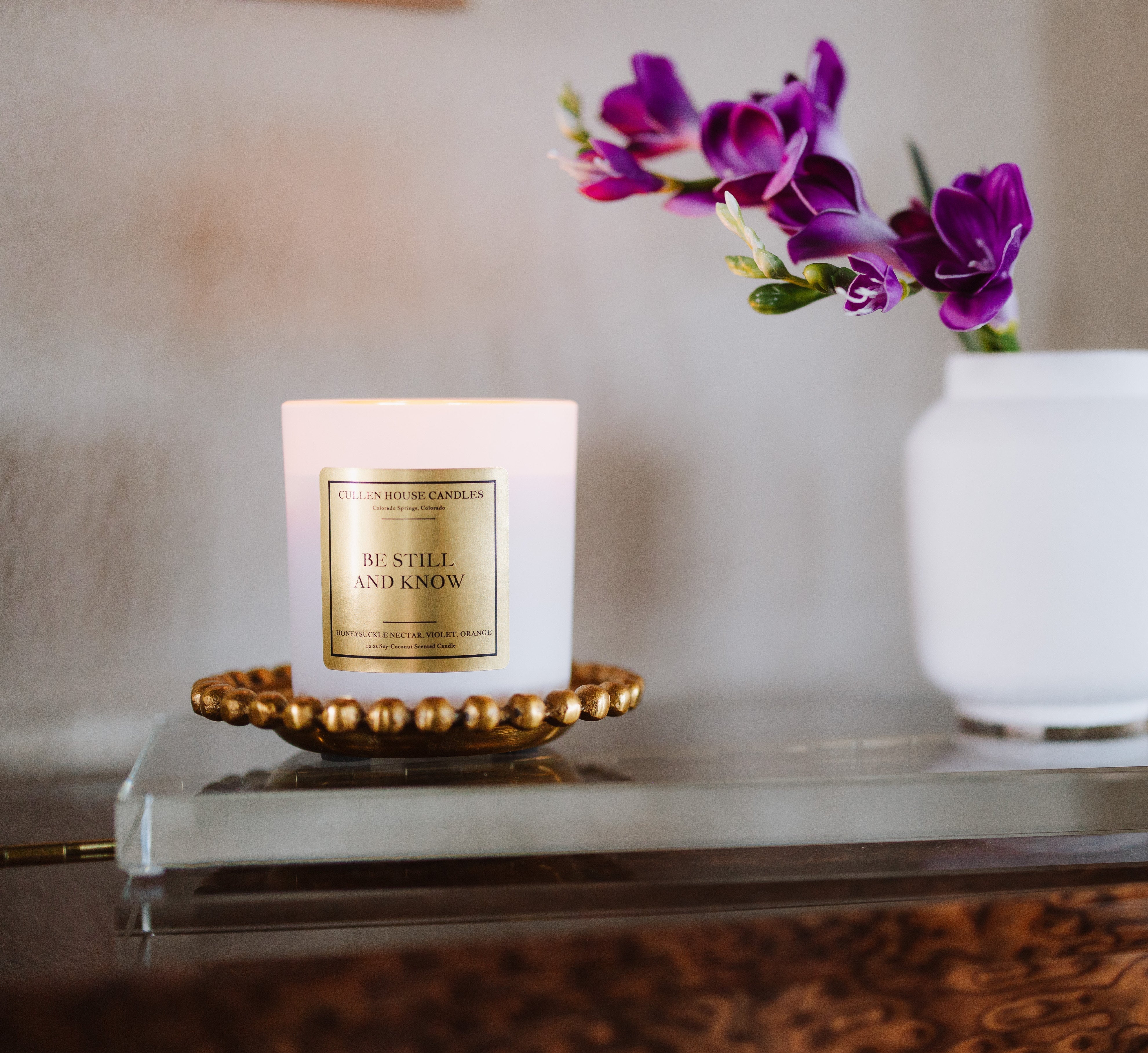 Candle with a gold label on a decorative stand next to a vase with purple flowers.