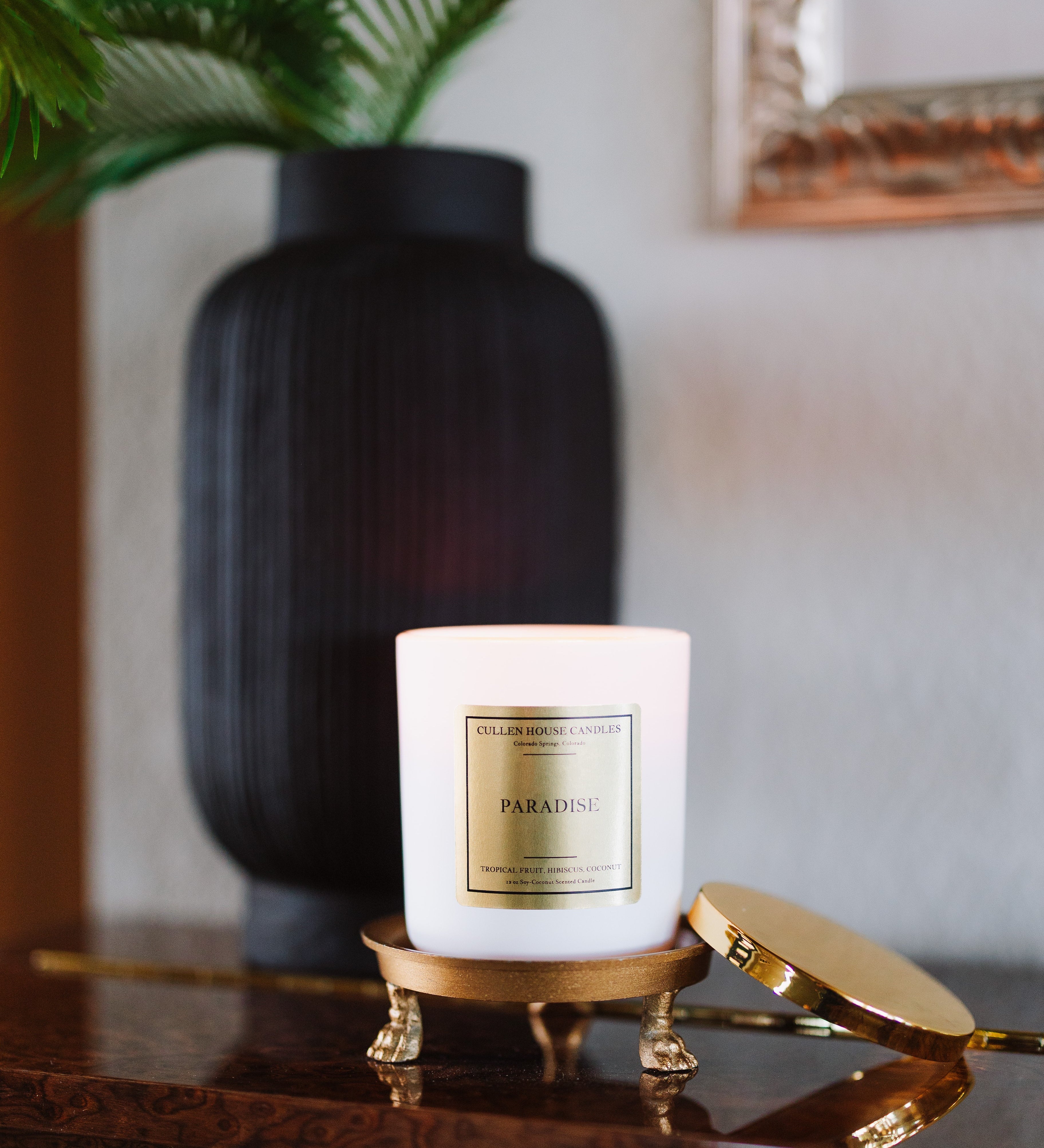 Candle labeled 'Paradise' on a decorative stand with a vase and plant in the background.