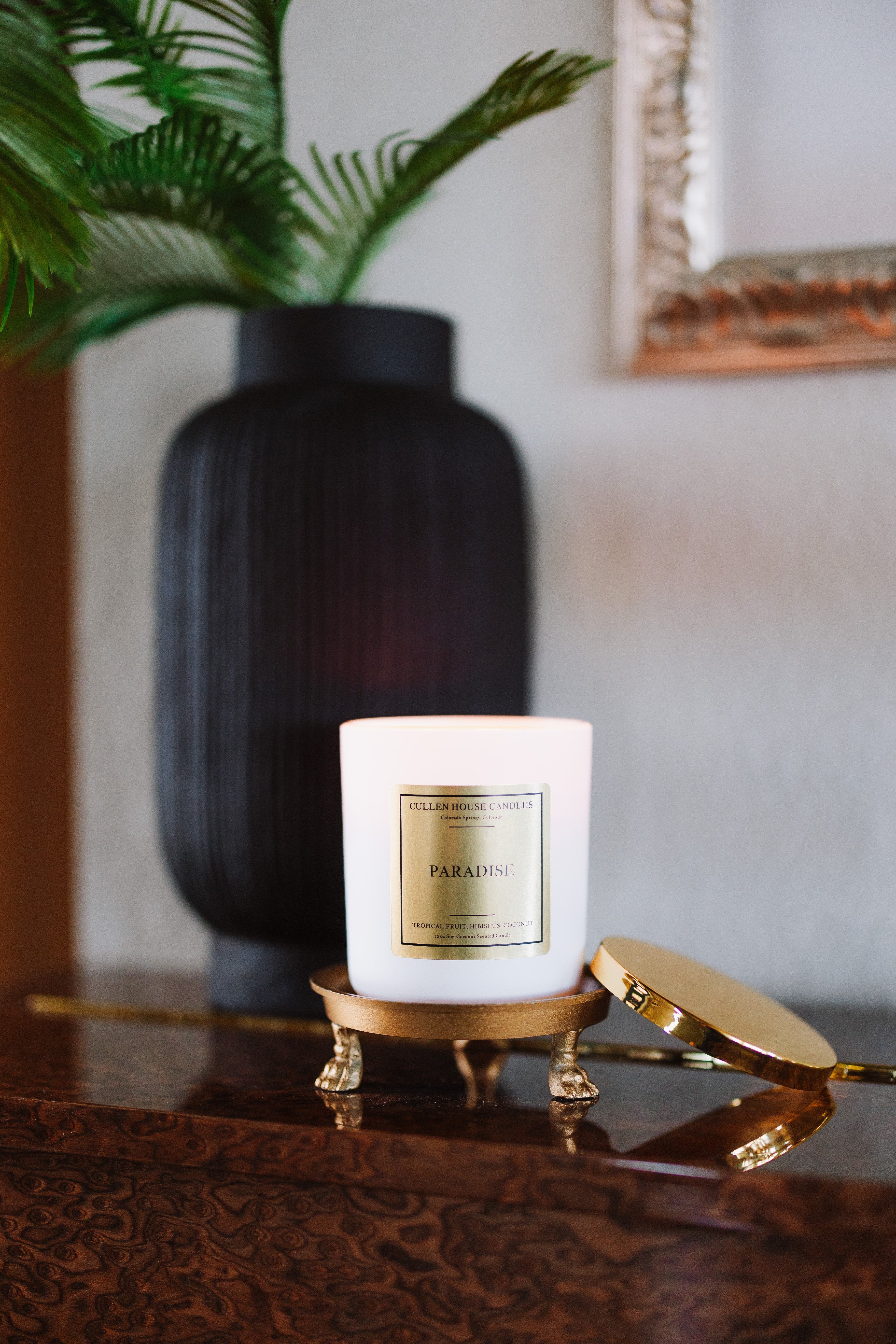 Candle labeled 'Paradise' on a decorative stand with a vase and plant in the background.