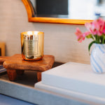 Candle on a wooden coaster with a vase of flowers and books in the background