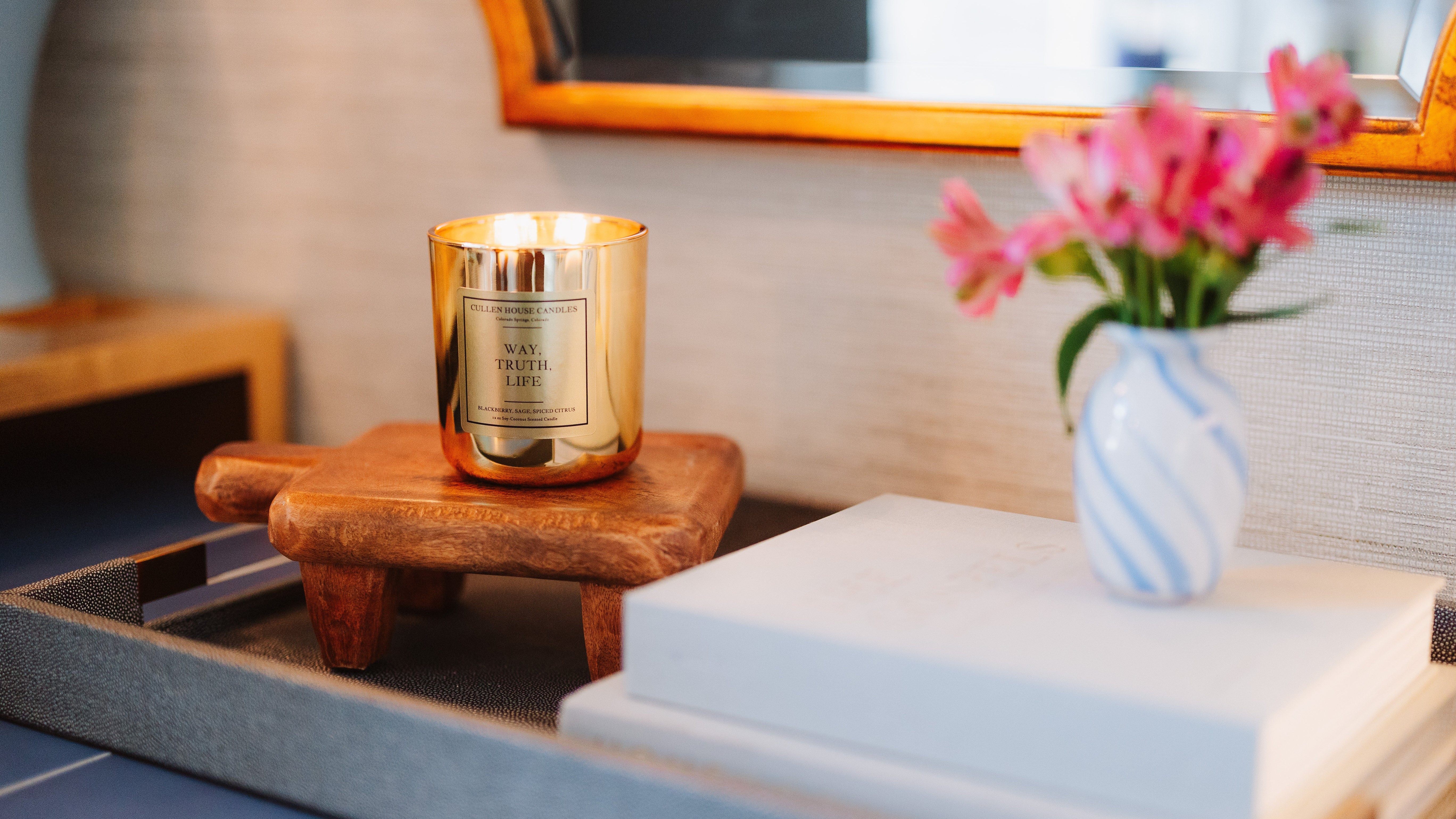Candle on a wooden coaster with a vase of flowers and books in the background