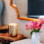 Candle with a gold holder on a wooden side table next to a vase with pink flowers.