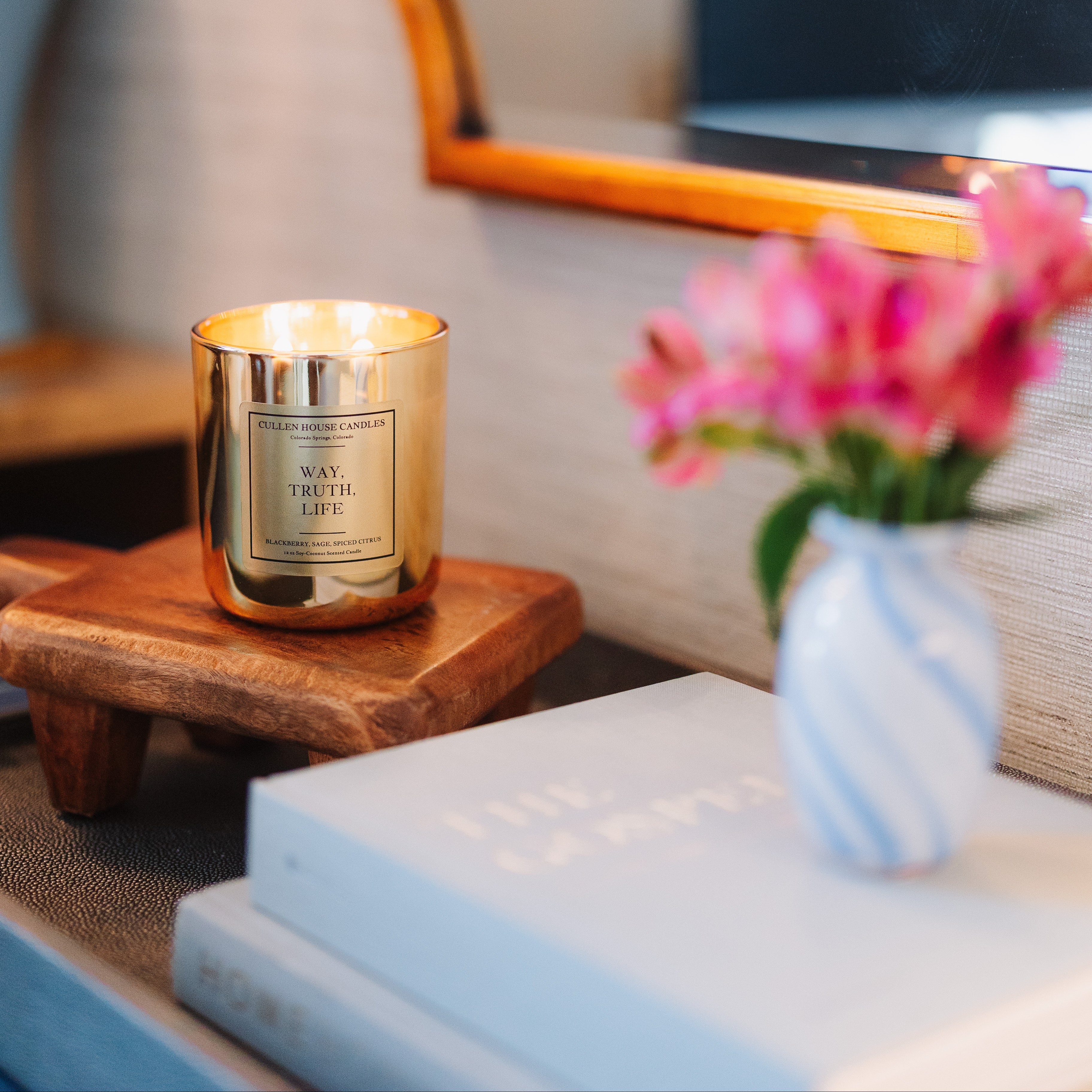 Candle with a gold holder on a wooden side table next to a vase with pink flowers.