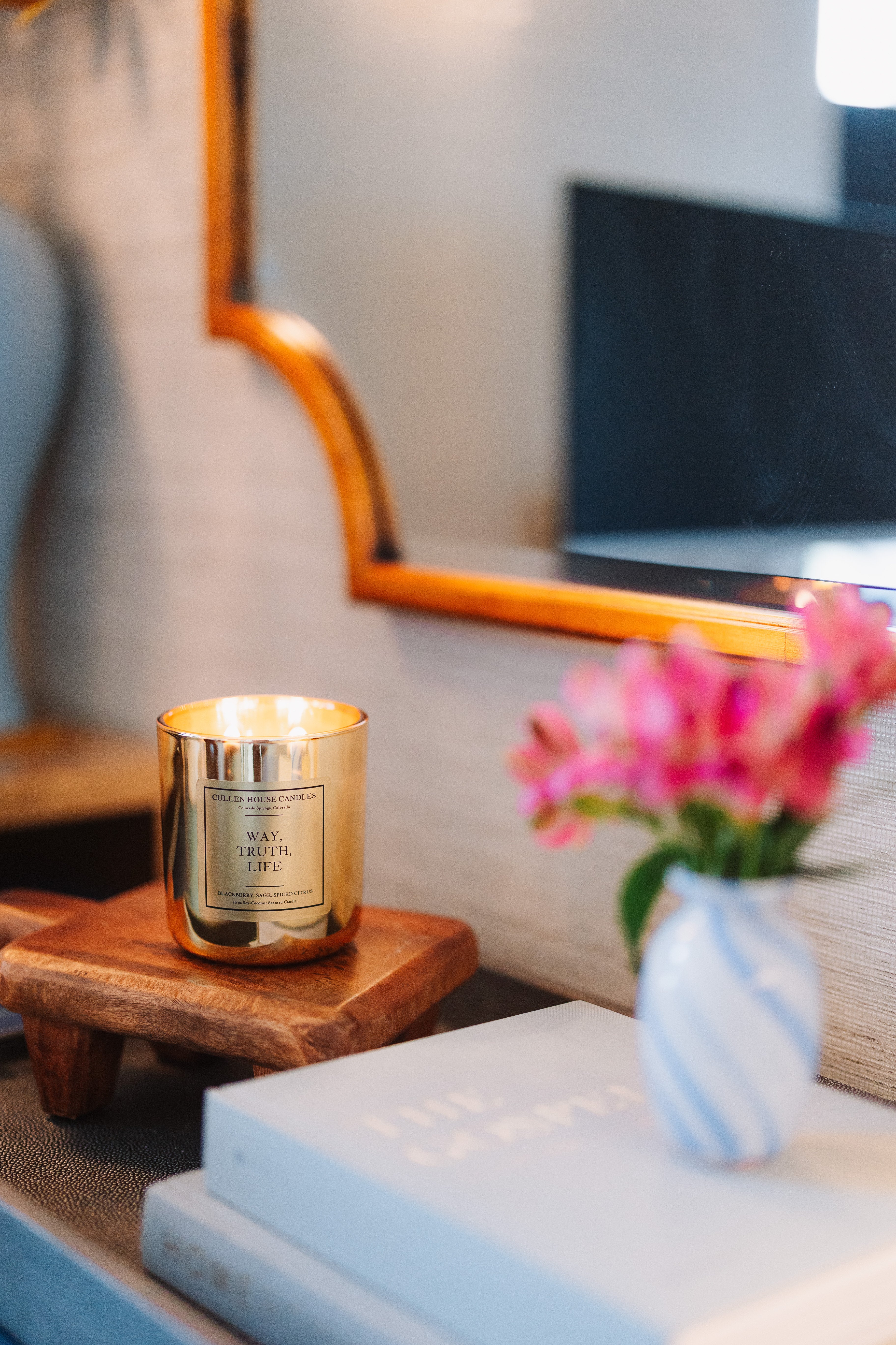 Candle with a gold holder on a wooden side table next to a vase with pink flowers.