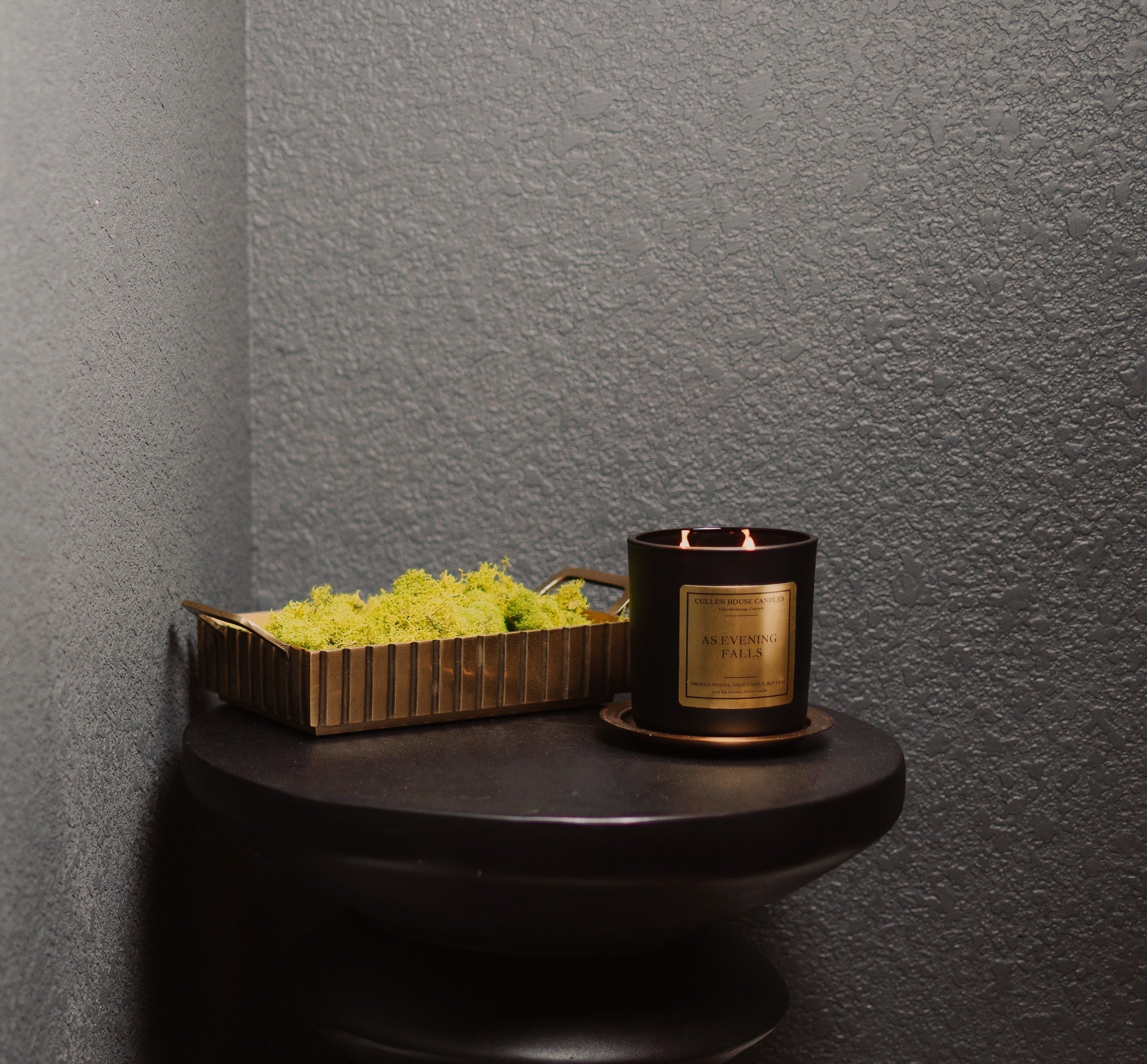 Black candle with gold label on a dark surface against a gray wall