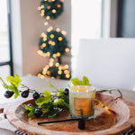 Candle on a wooden tray with decorative branches and blurred lights in the background
