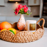 Wicker tray with fruits, a candle, and a vase with flowers on a kitchen counter.