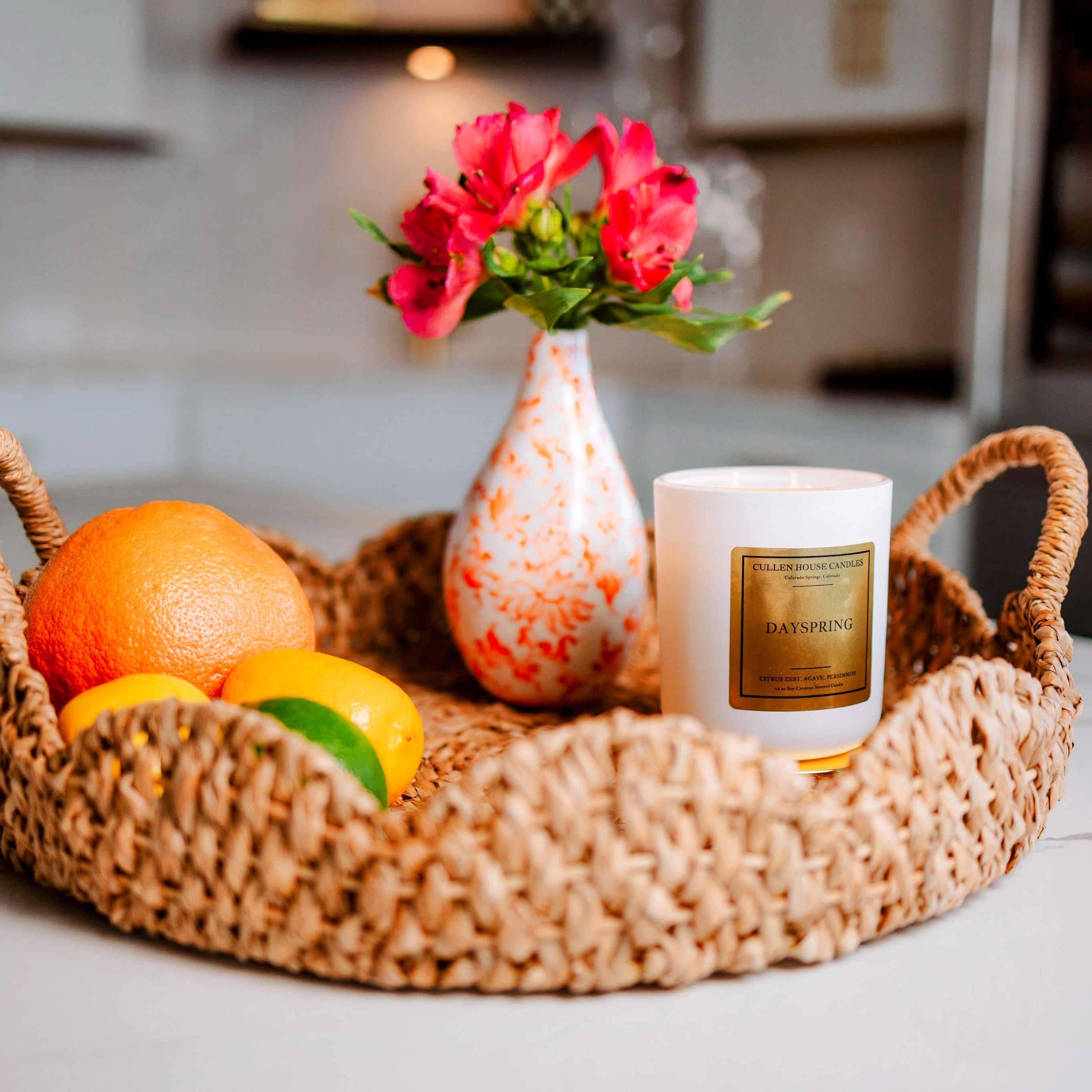 Wicker tray with fruits, a candle, and a vase with flowers on a kitchen counter.