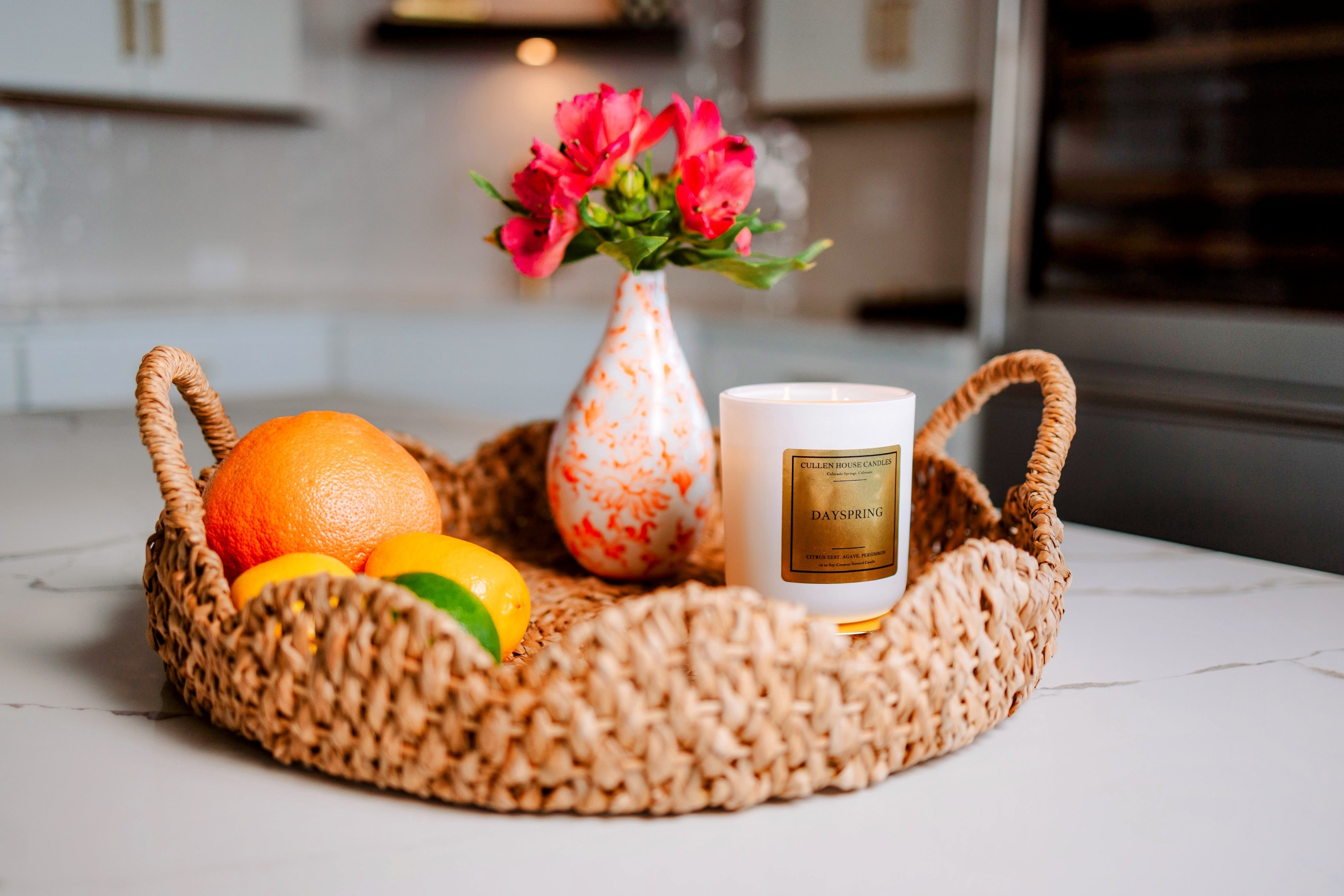 Wicker tray with fruits, a candle, and a vase with flowers on a kitchen counter.