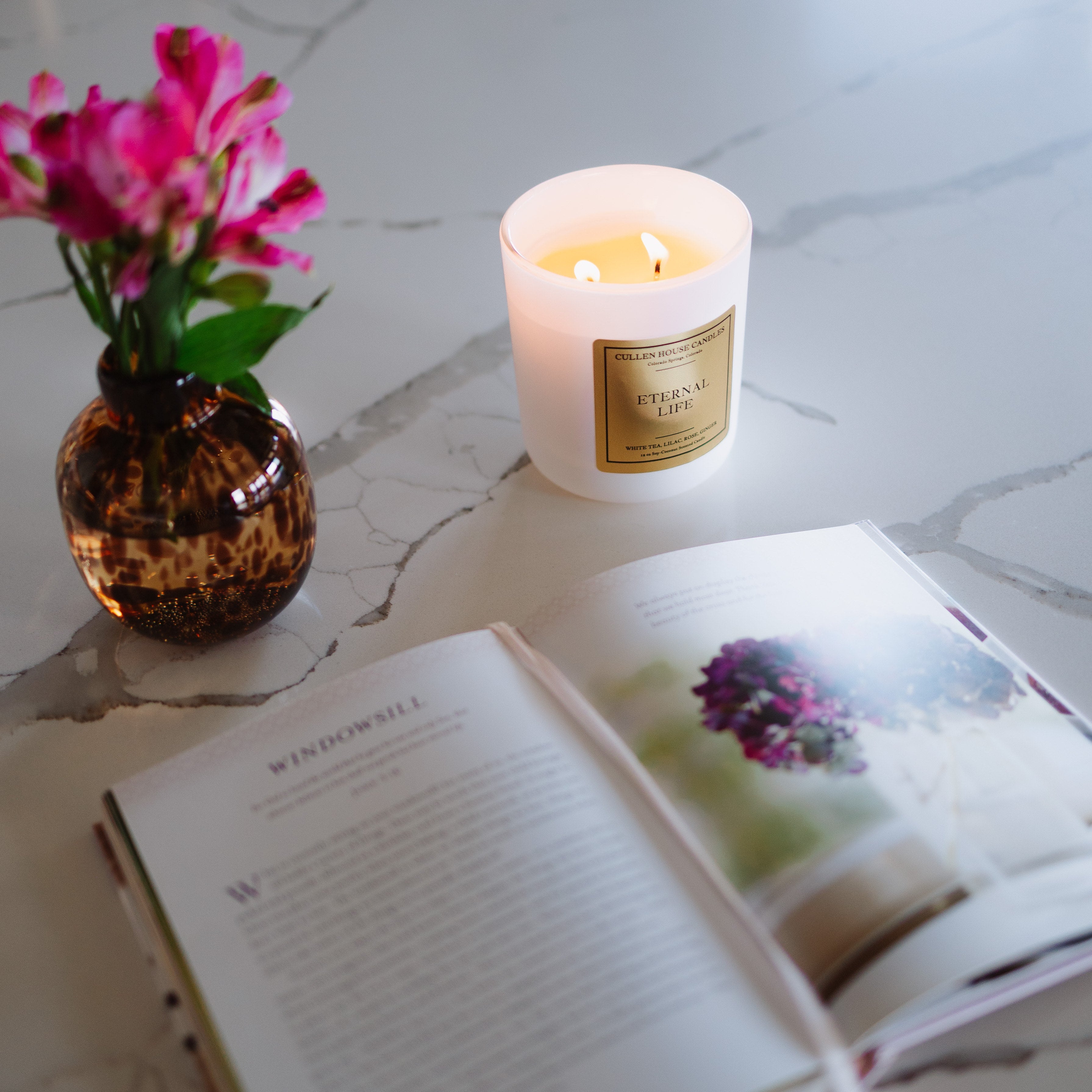 Candle, vase with flowers, and open book on a marble surface