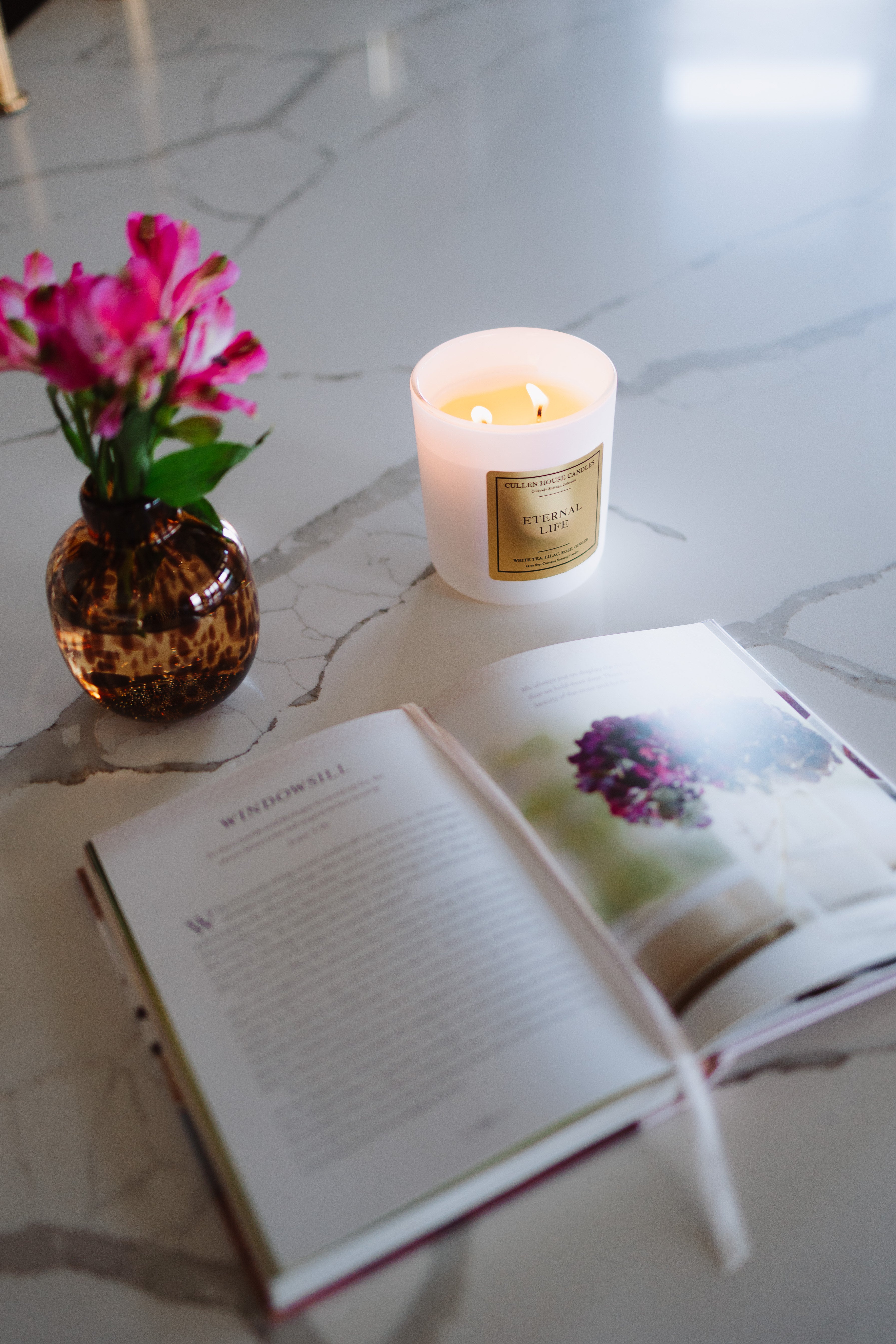 Candle, vase with flowers, and open book on a marble surface