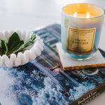 Candle labeled 'Green Pastures' on a magazine with a white bowl containing leaves.
