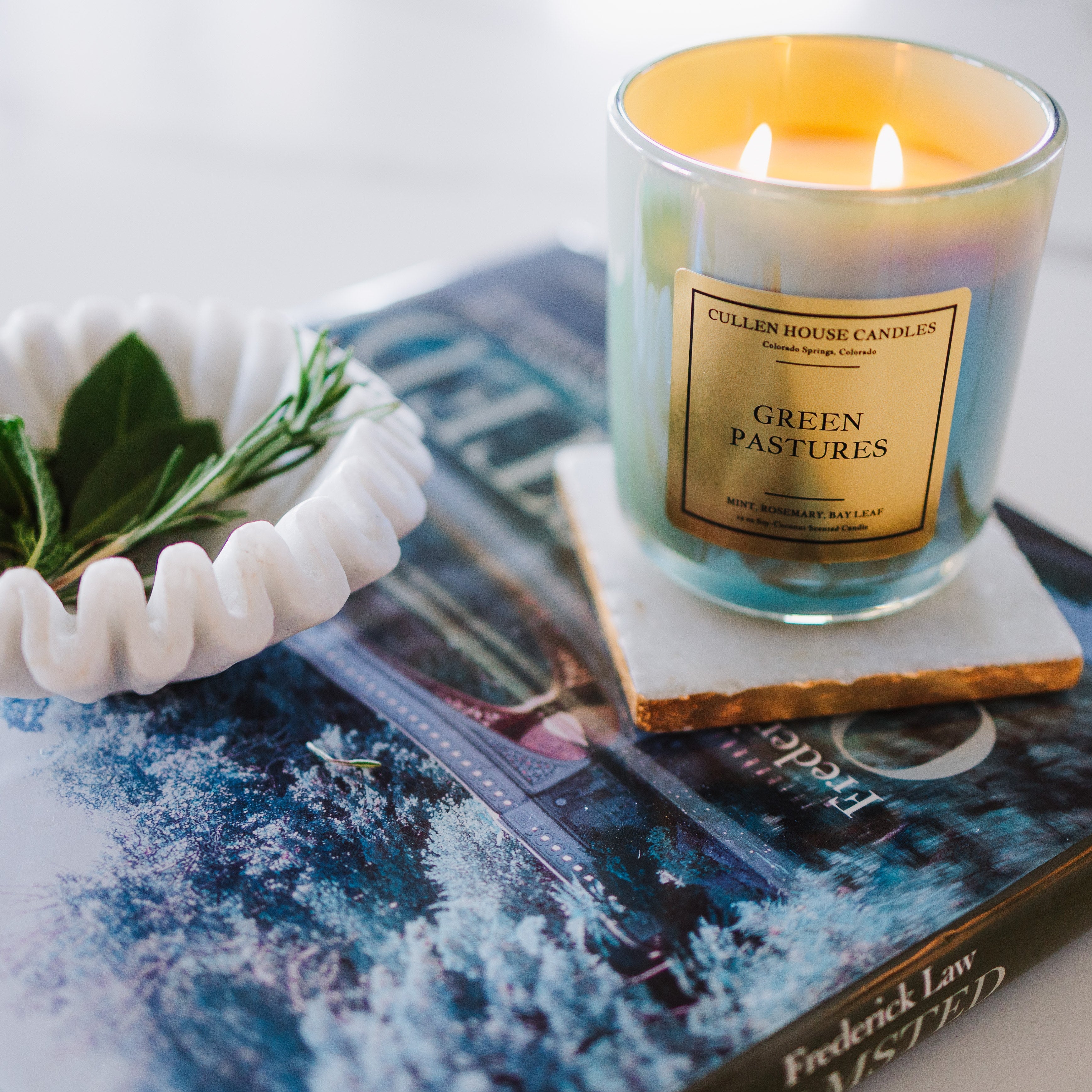 Candle labeled 'Green Pastures' on a magazine with a white bowl containing leaves.