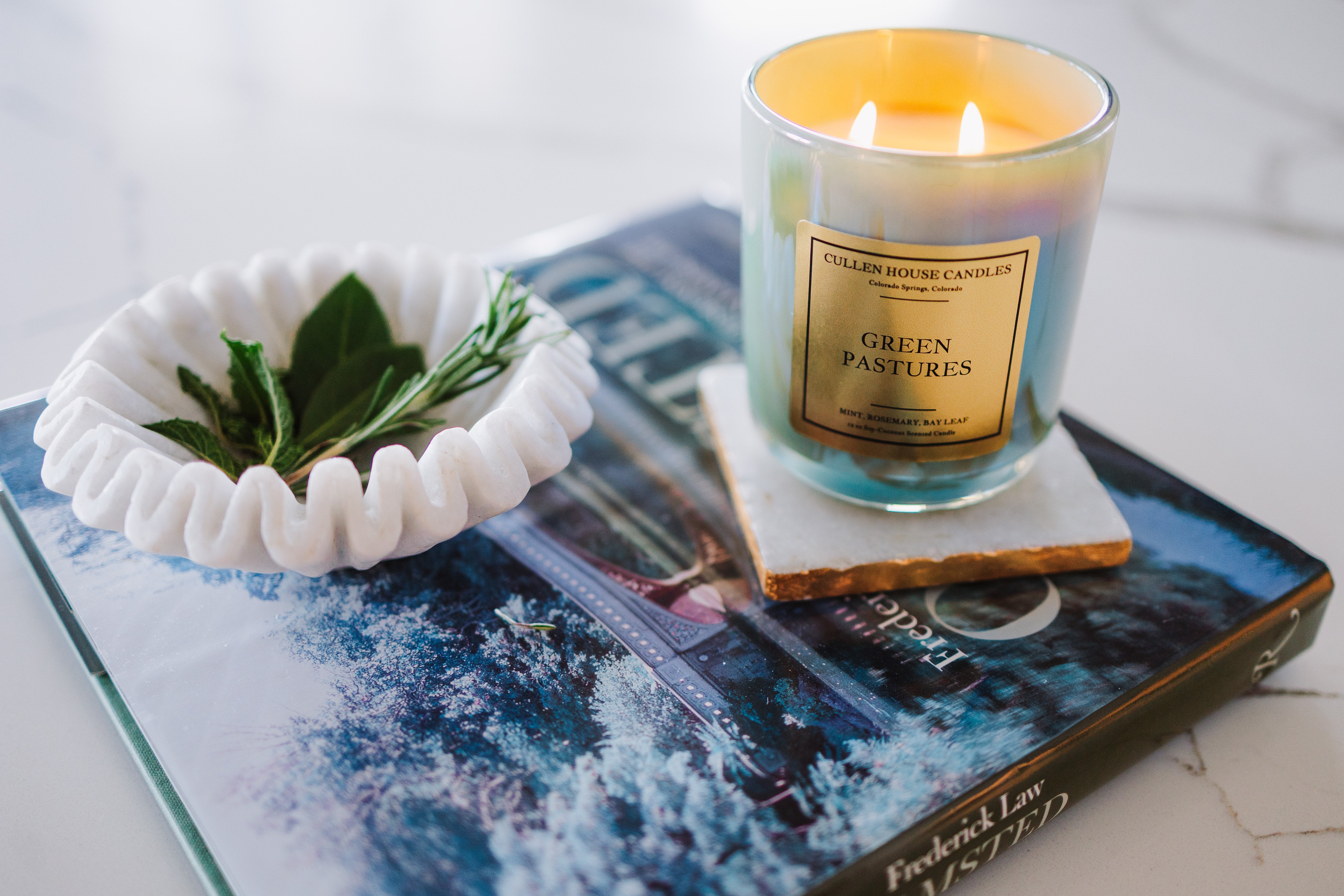 Candle labeled 'Green Pastures' on a magazine with a white bowl containing leaves.