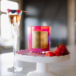 Candle labeled 'Banquet of Love' on a white stand with a glass of champagne and berries in the background.