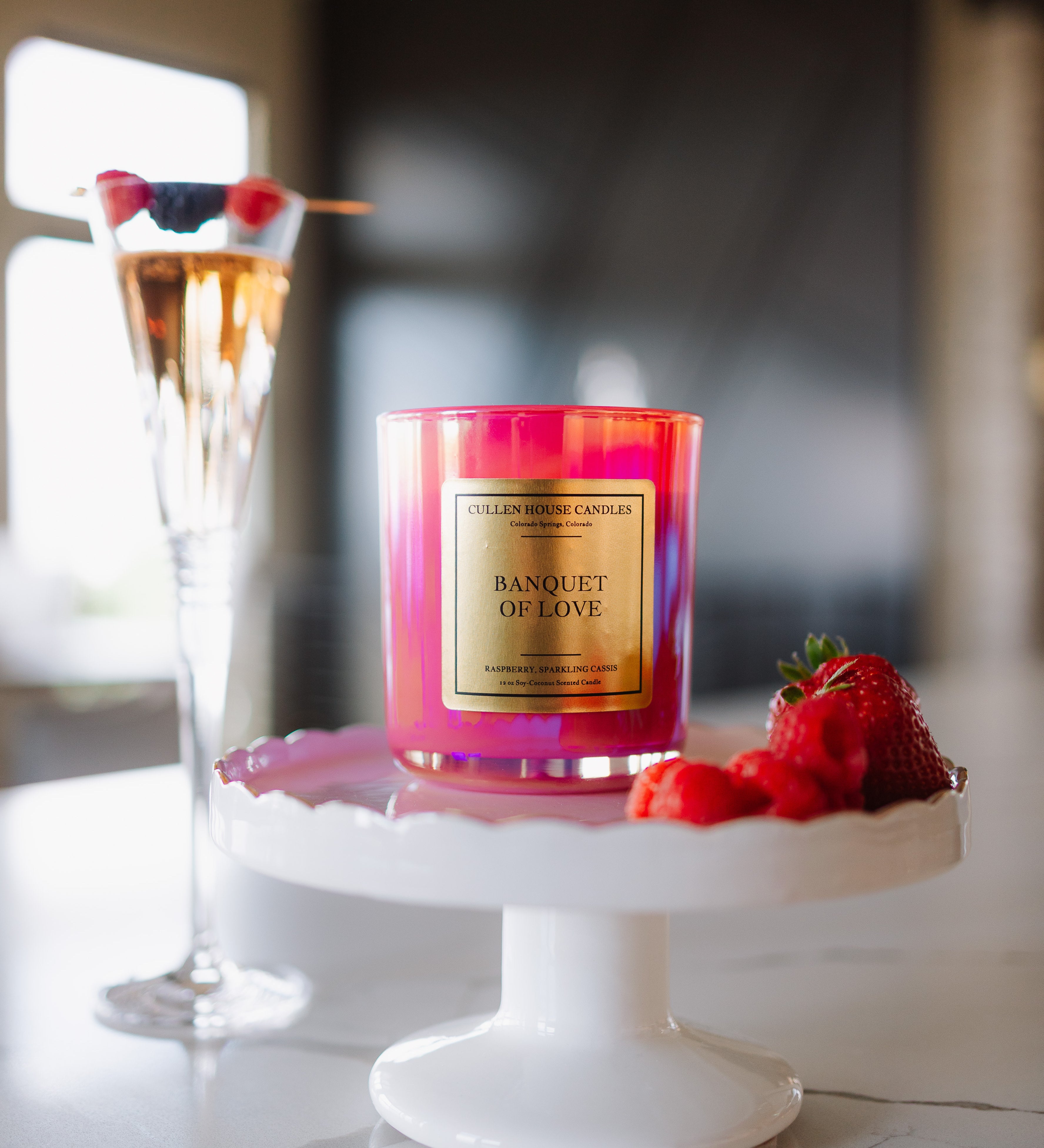 Candle labeled 'Banquet of Love' on a white stand with a glass of champagne and berries in the background.