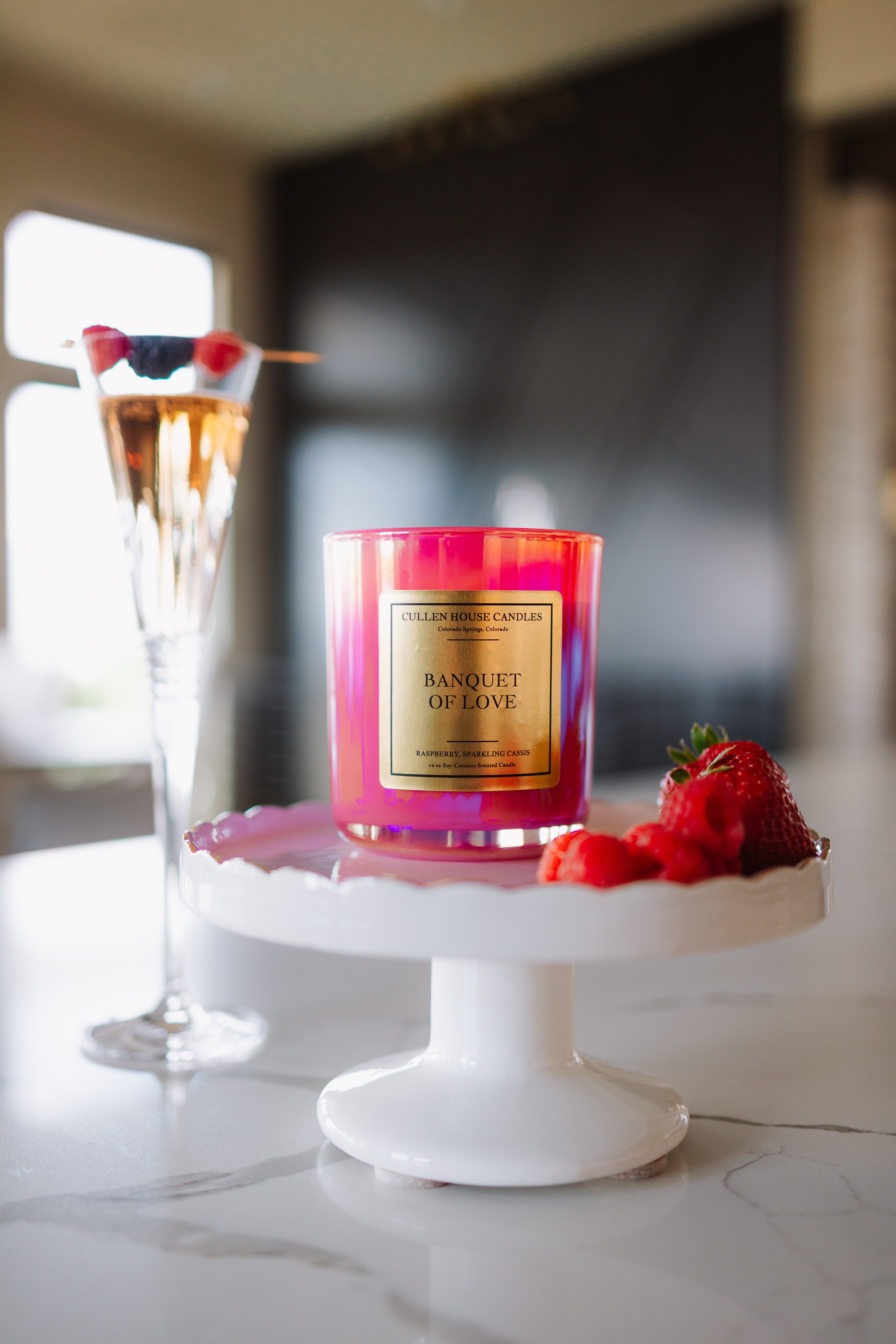 Candle labeled 'Banquet of Love' on a white stand with a glass of champagne and berries in the background.