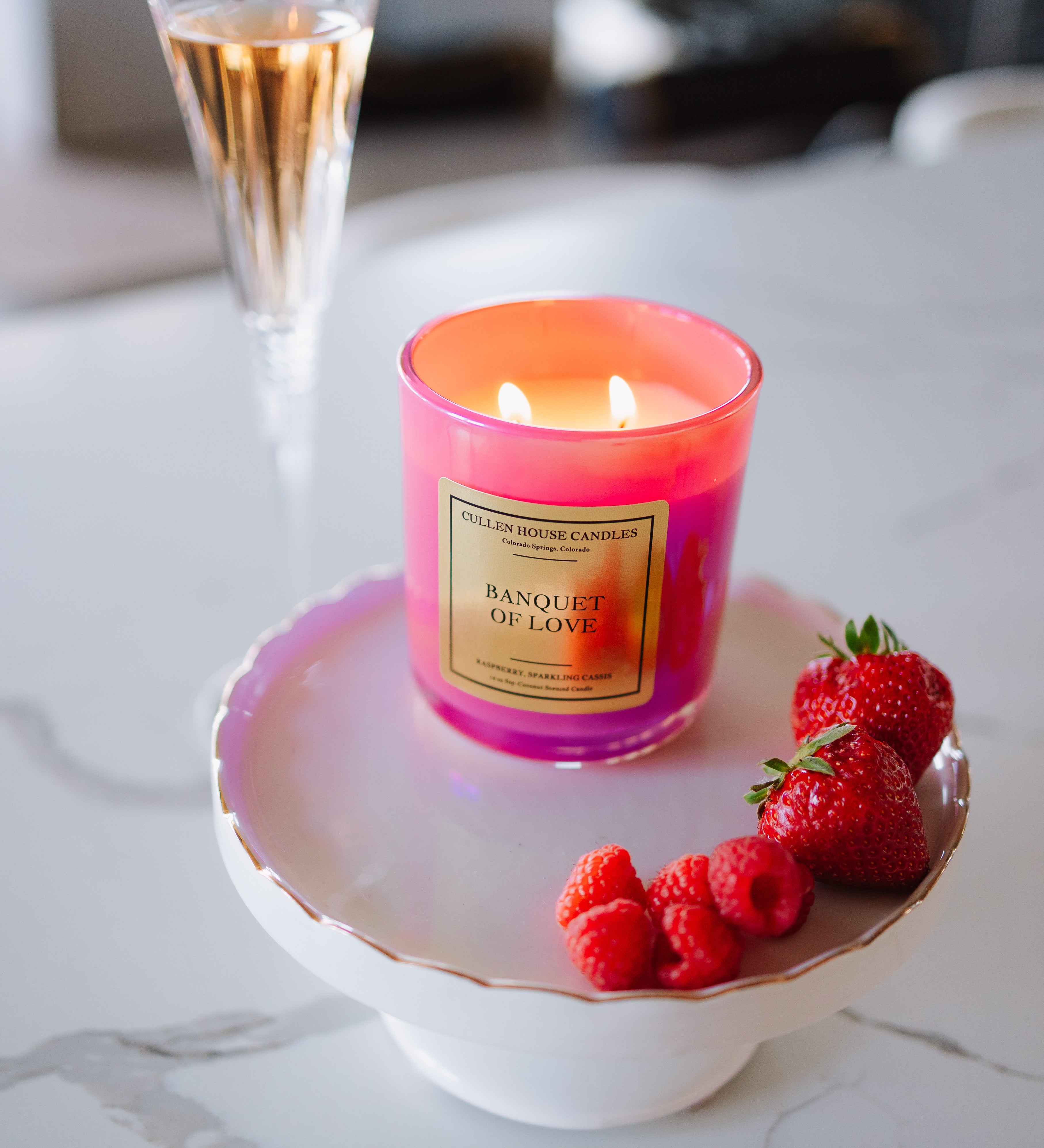 Candle labeled 'Banquet of Love' on a white dish with raspberries and strawberries, next to a cocktail.