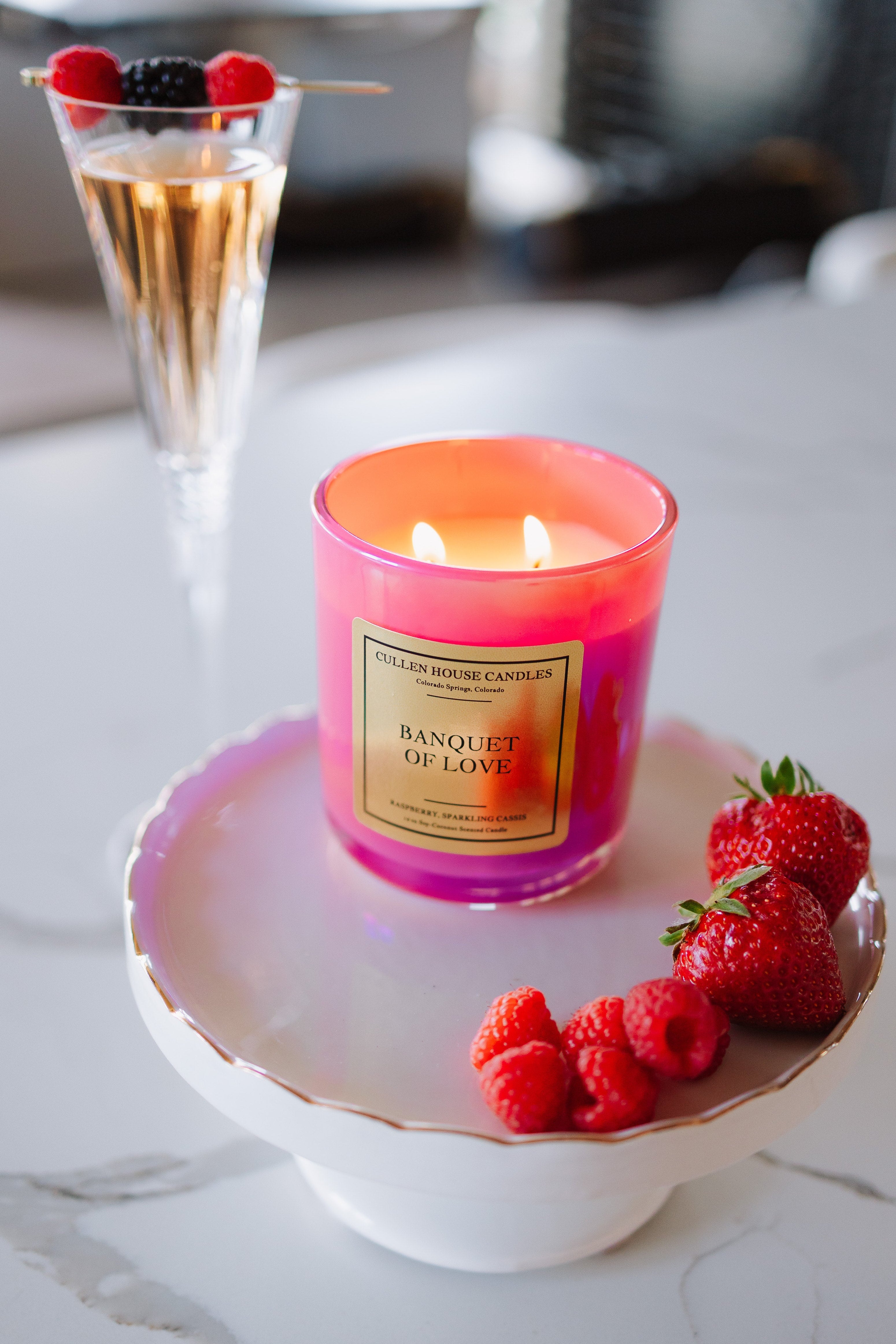 Candle labeled 'Banquet of Love' on a white dish with raspberries and strawberries, next to a cocktail.