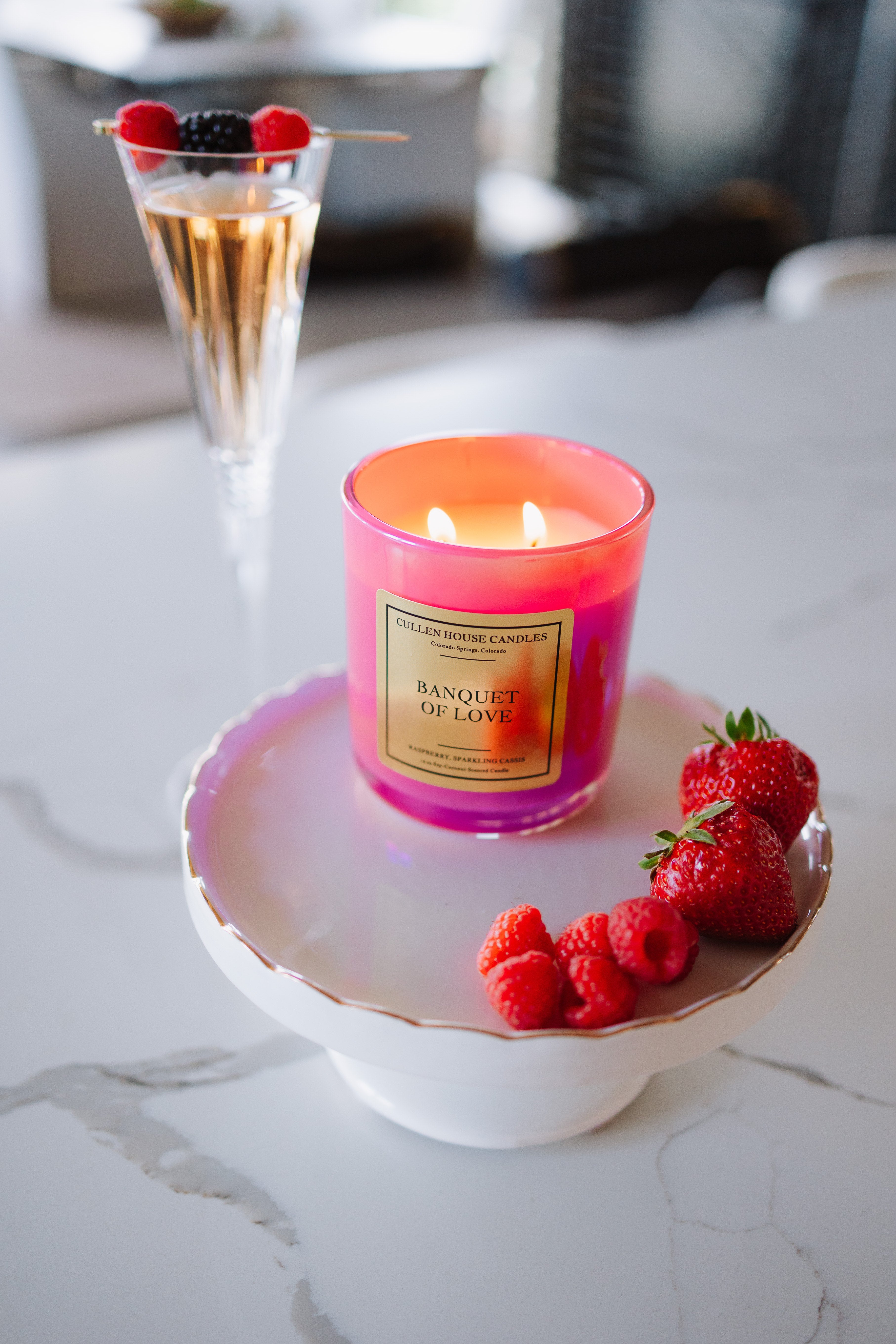 Candle labeled 'Banquet of Love' on a white dish with raspberries and strawberries, next to a cocktail.