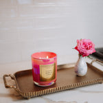 Candle with a pink and red design on a gold tray with a vase of pink flowers on a marble surface.