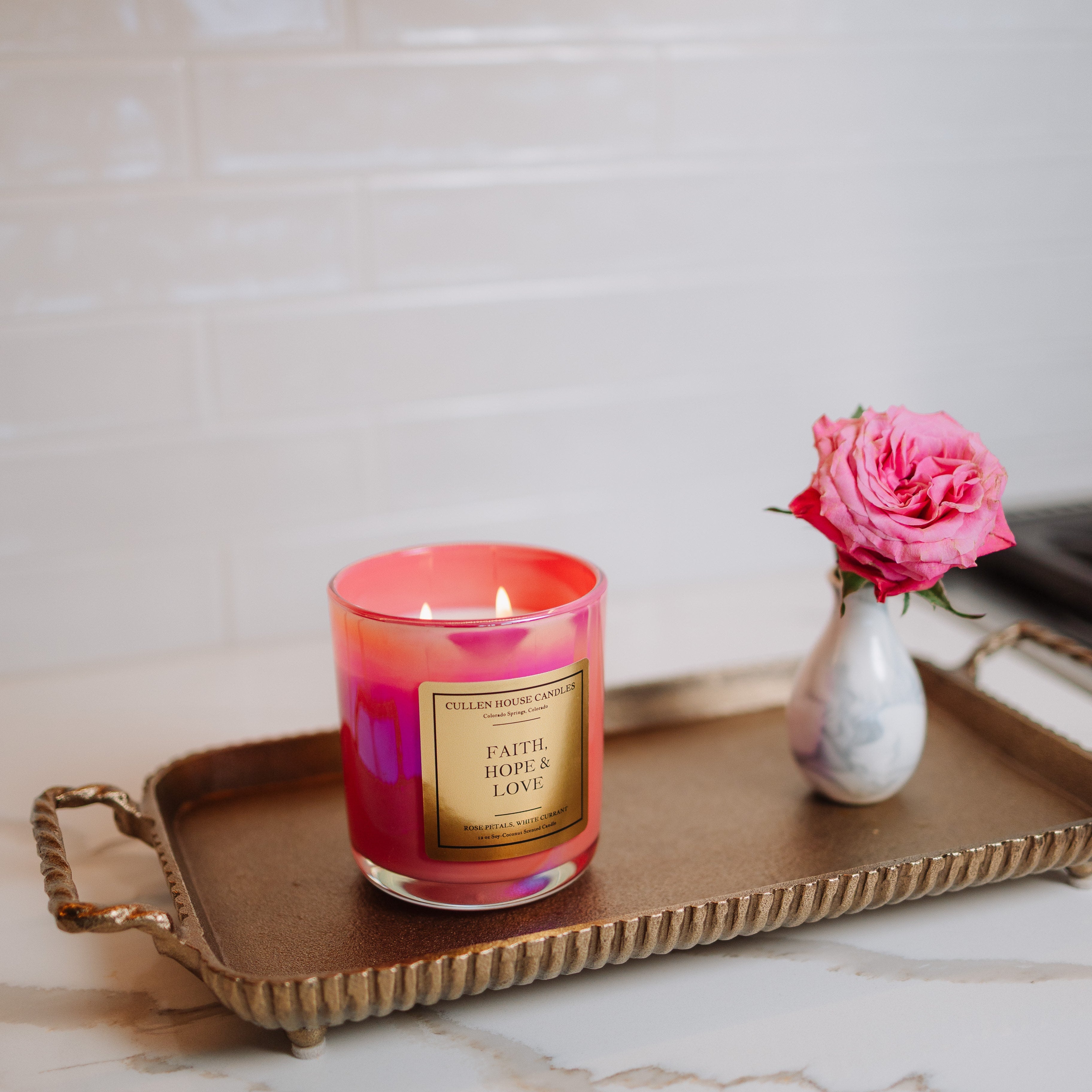 Candle with a pink and red design on a gold tray with a vase of pink flowers on a marble surface.