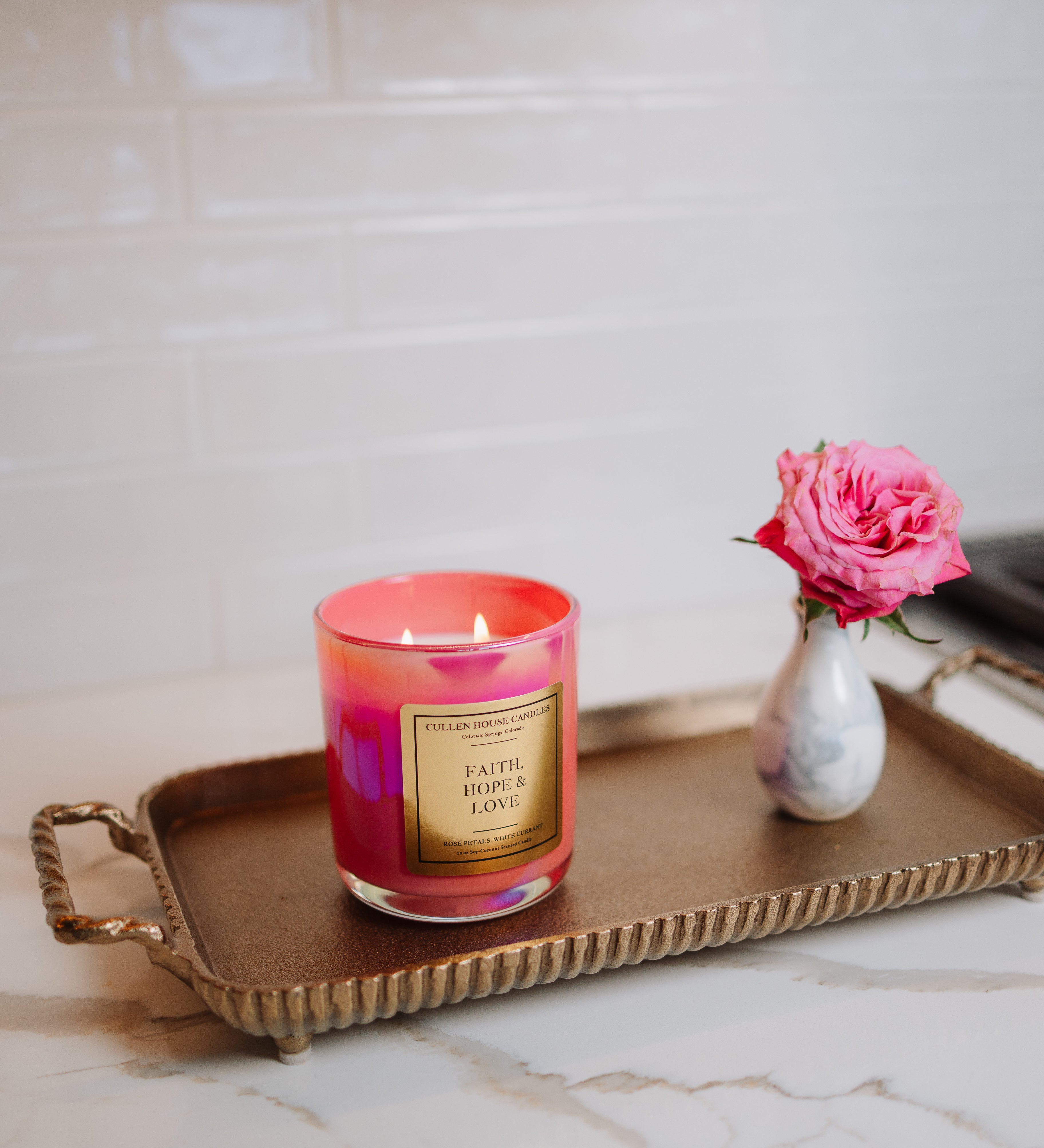 Candle with a pink and red design on a gold tray with a vase of pink flowers on a marble surface.