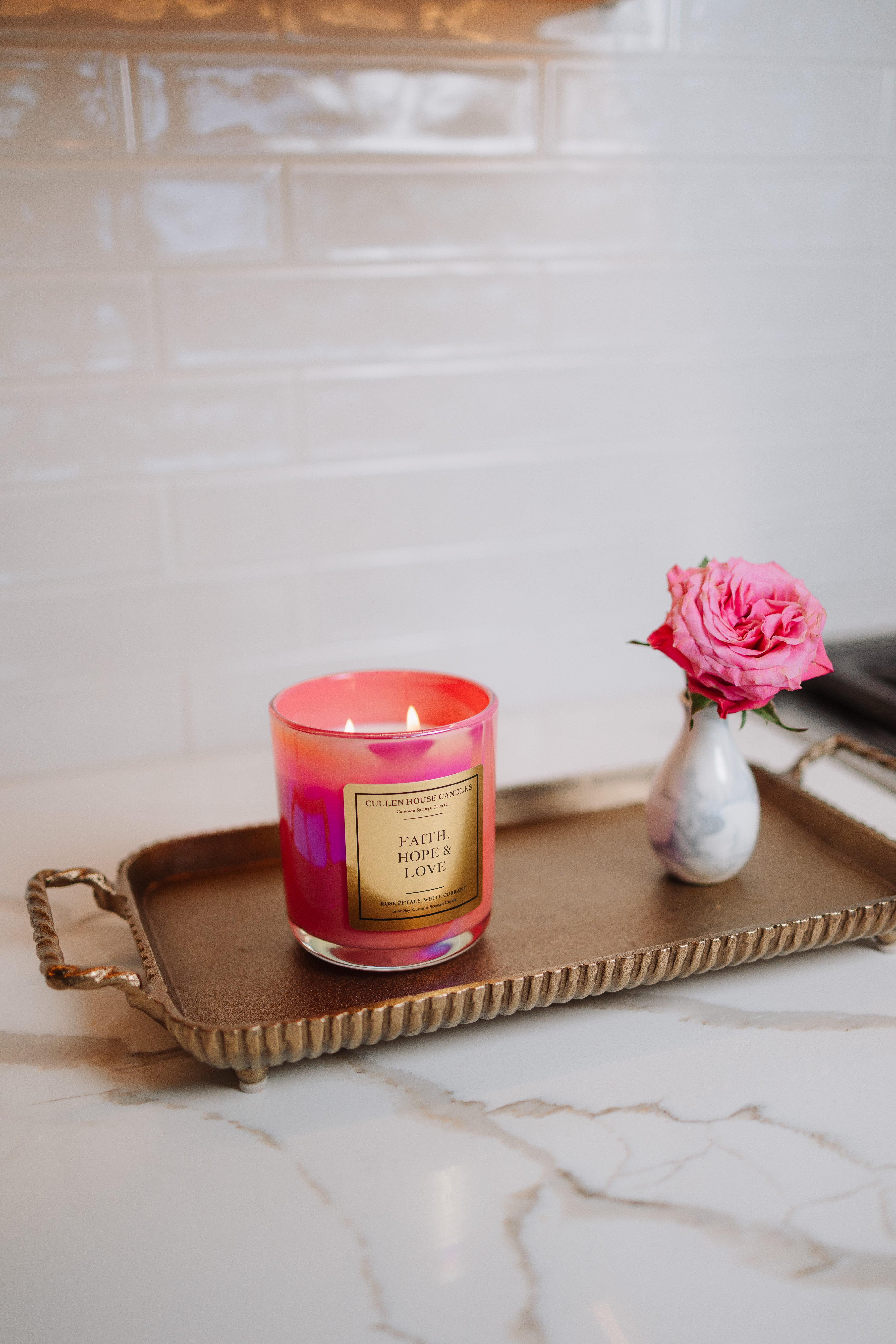 Candle with a pink and red design on a gold tray with a vase of pink flowers on a marble surface.
