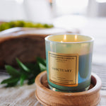 Candle labeled 'Sanctuary' on a wooden coaster with a blurred indoor background