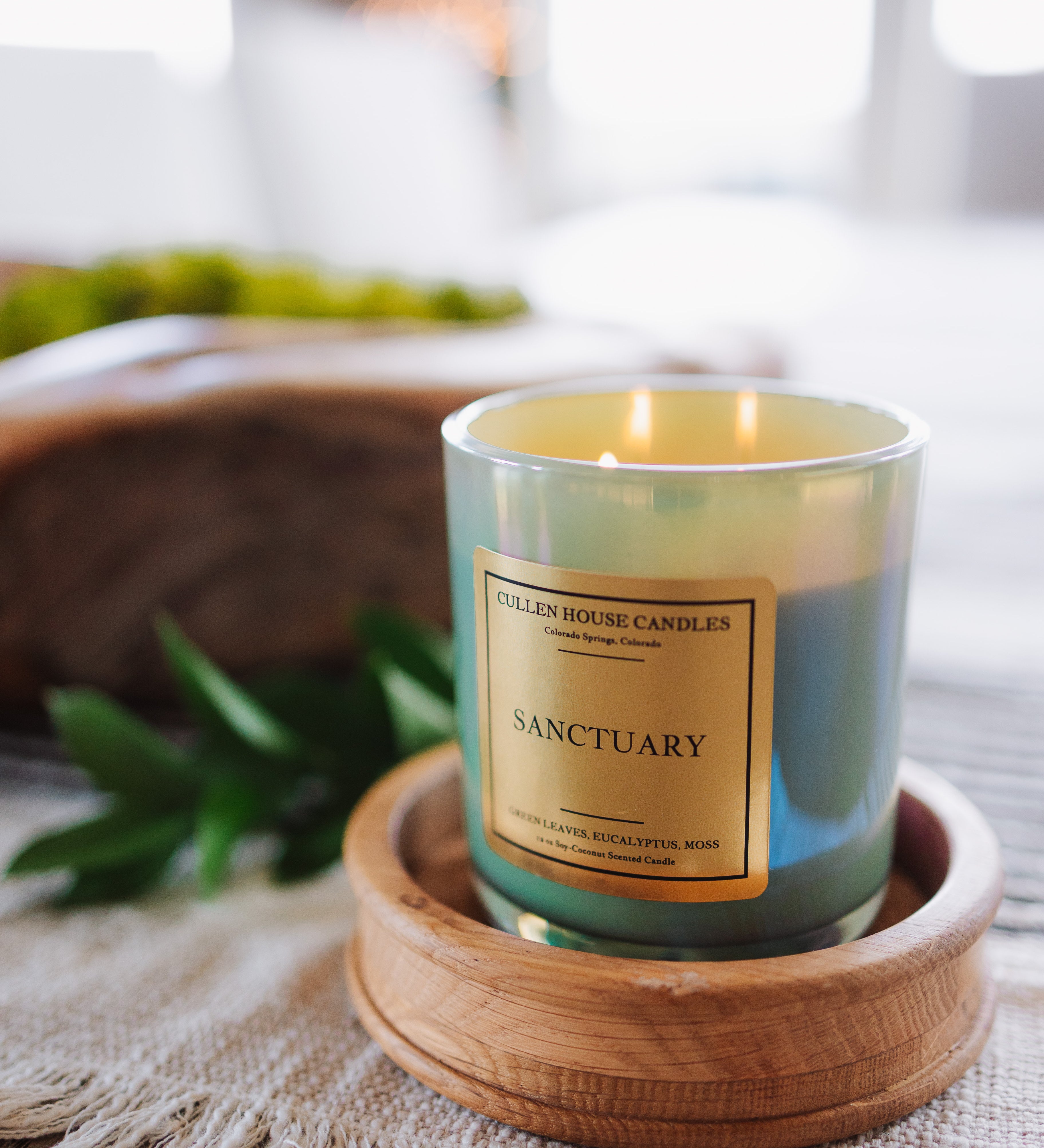 Candle labeled 'Sanctuary' on a wooden coaster with a blurred indoor background