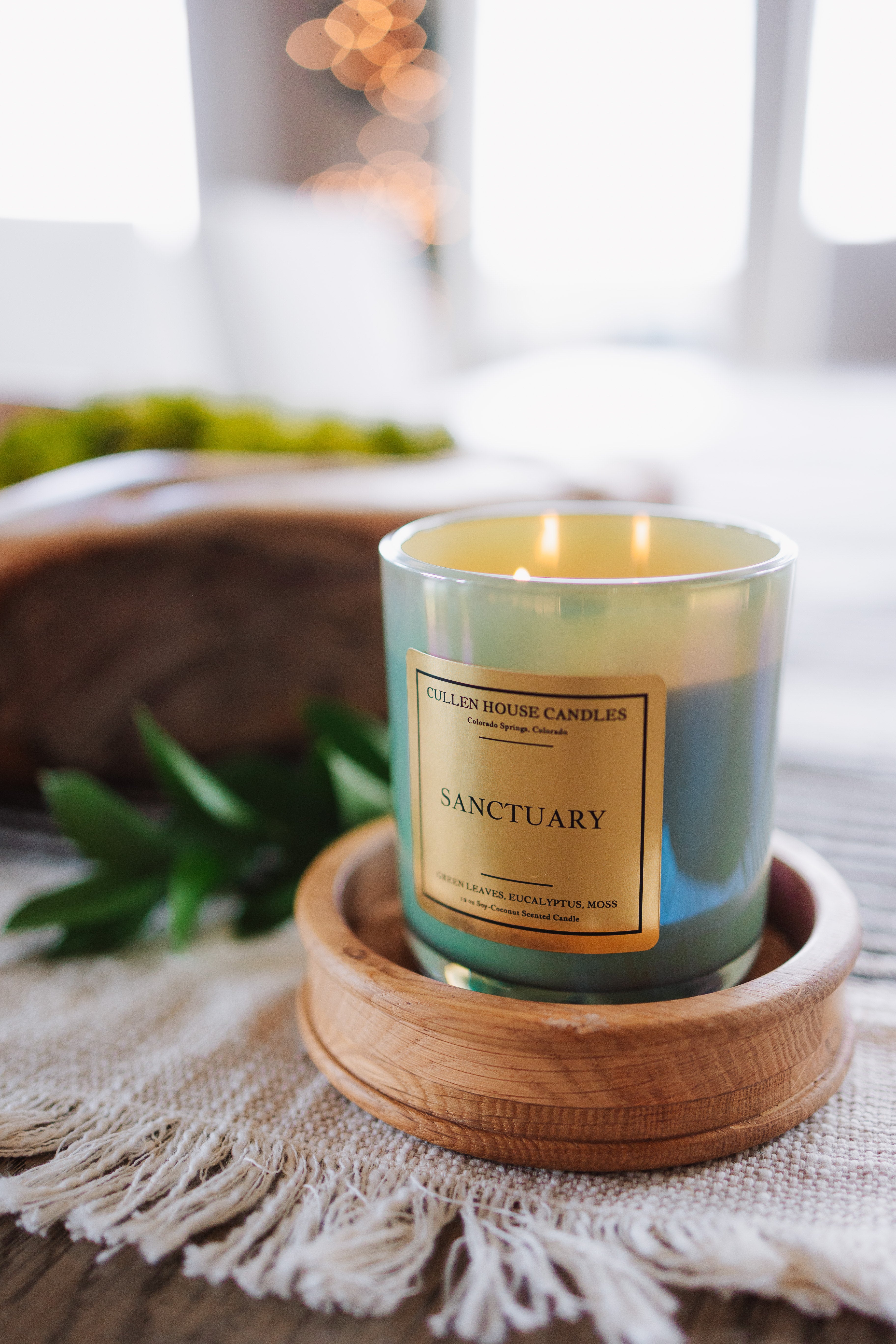 Candle labeled 'Sanctuary' on a wooden coaster with a blurred indoor background