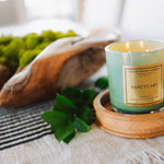 Candle labeled 'Sanctuary' on a wooden coaster with greenery and a woven basket in the background.