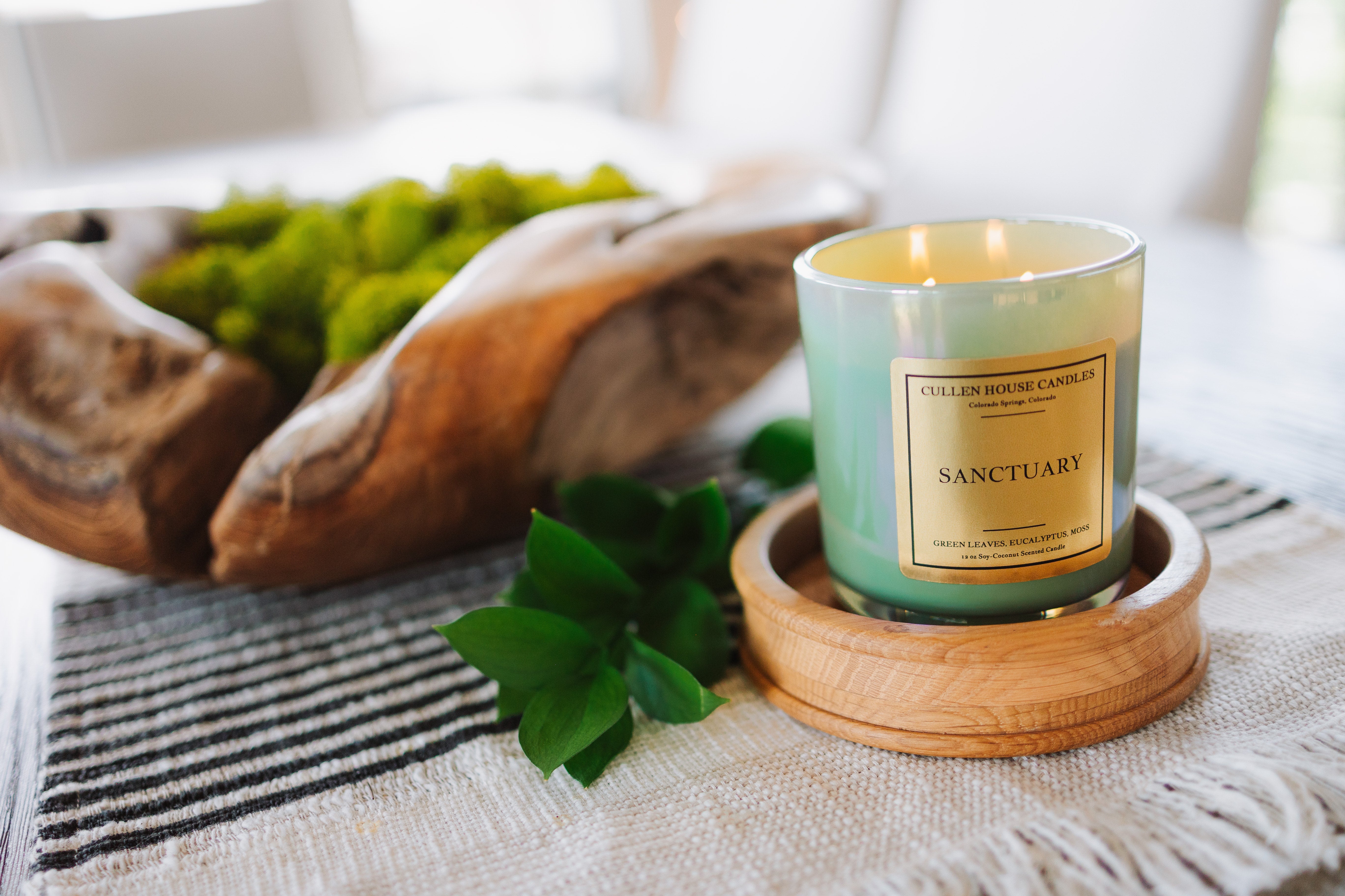 Candle labeled 'Sanctuary' on a wooden coaster with greenery and a woven basket in the background.