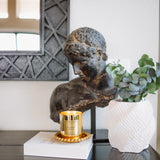 Decorative setup with a bronze bust, gold candle, and white vase on a dark surface.