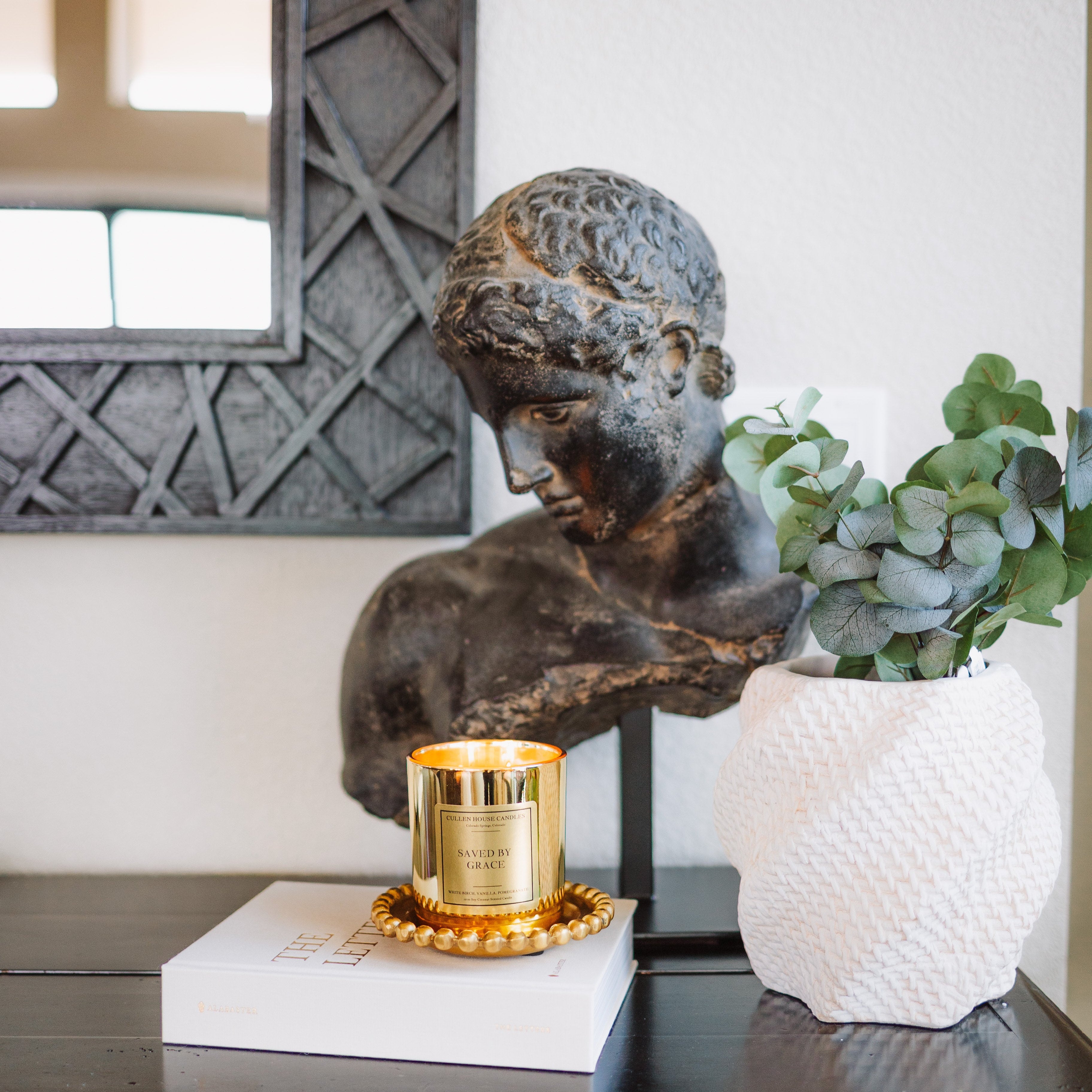 Decorative setup with a bronze bust, gold candle, and white vase on a dark surface.