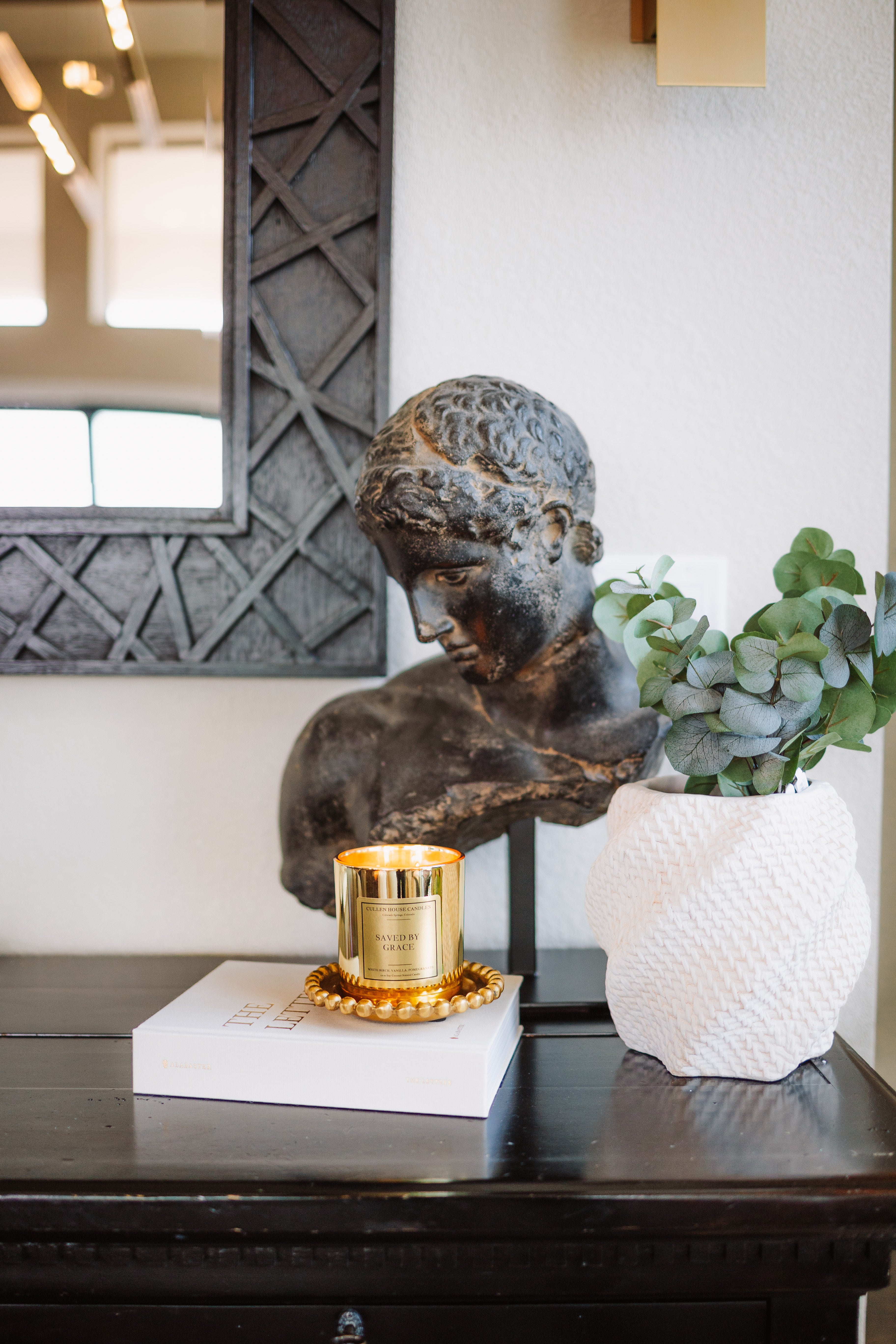 Decorative setup with a bronze bust, gold candle, and white vase on a dark surface.