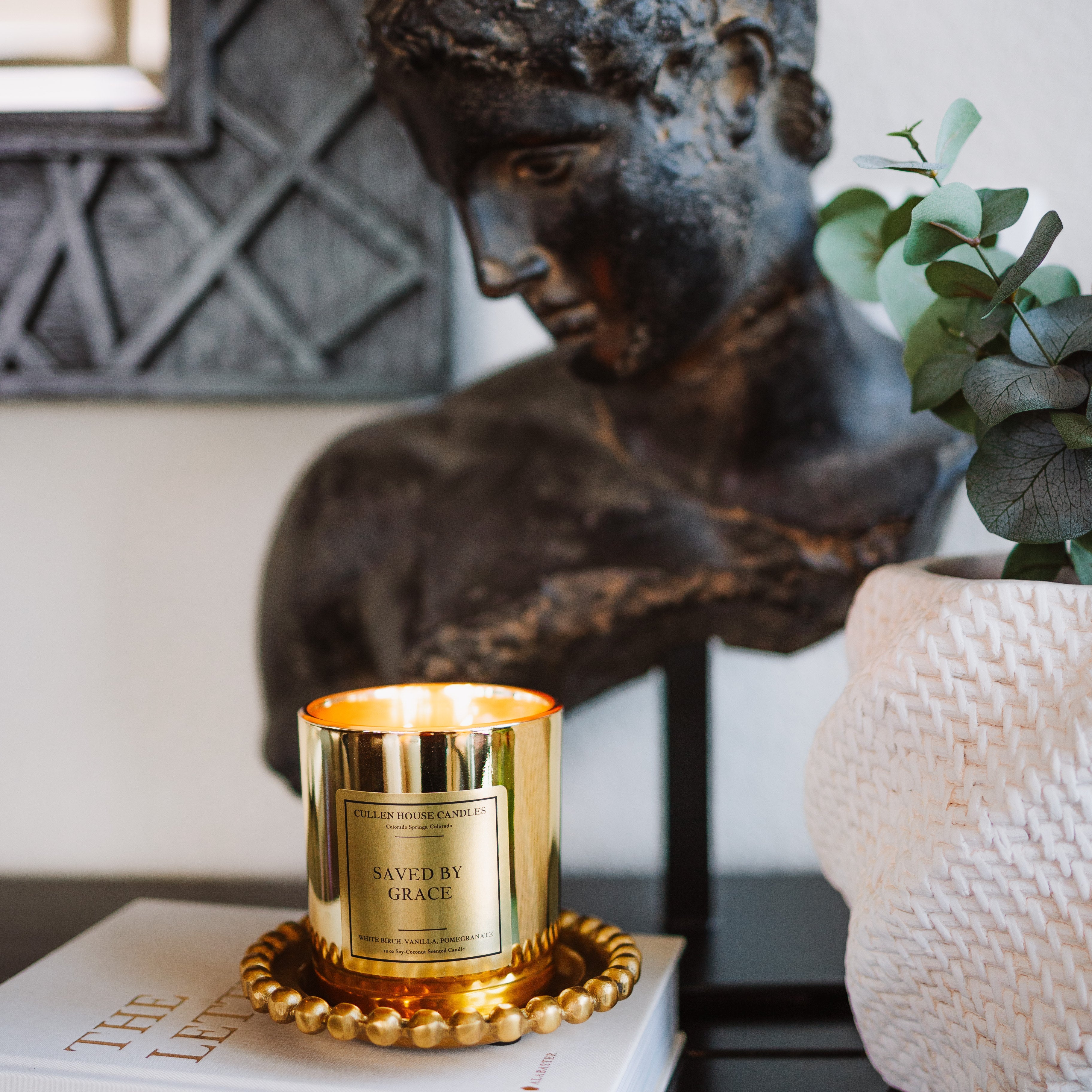 Candle with decorative label on a book next to a classical statue and plant