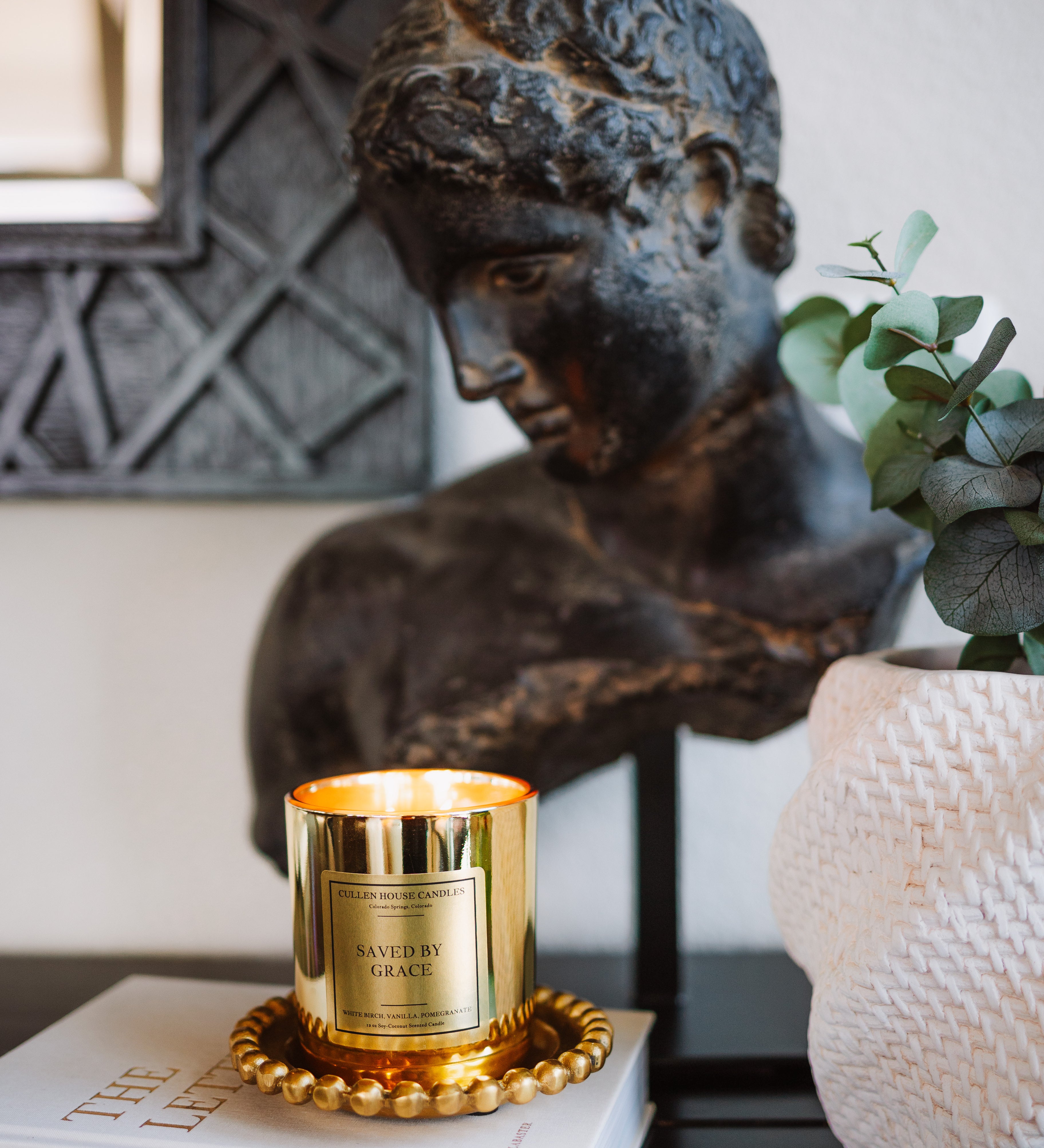 Candle with decorative label on a book next to a classical statue and plant