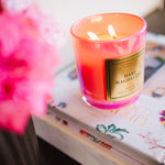 Candle with a visible brand label on a surface with flowers and books in the background
