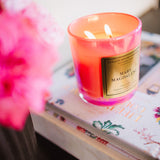 Candle with a visible brand label on a surface with flowers and books in the background