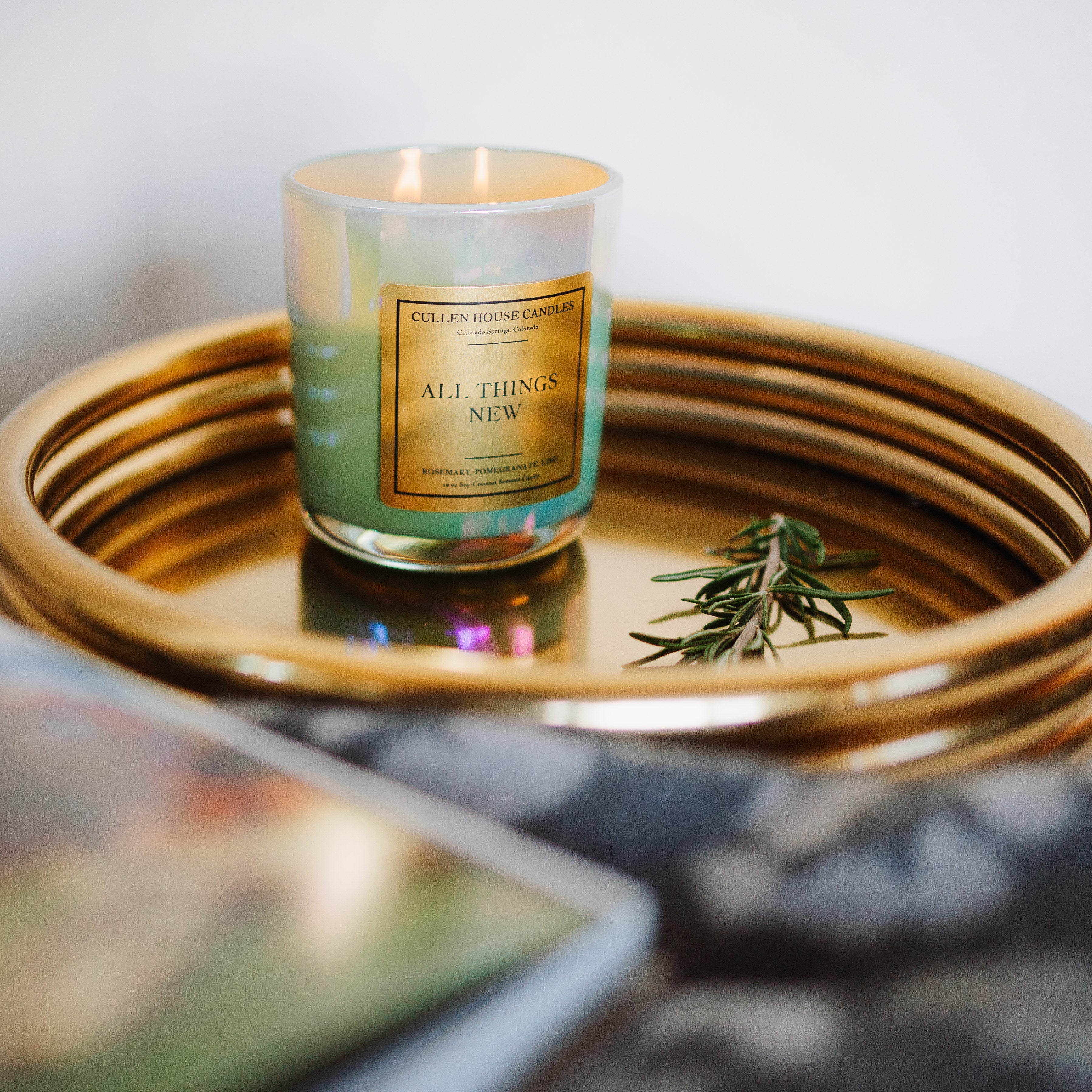 Candle labeled 'All Things New' on a gold tray with a blurred background
