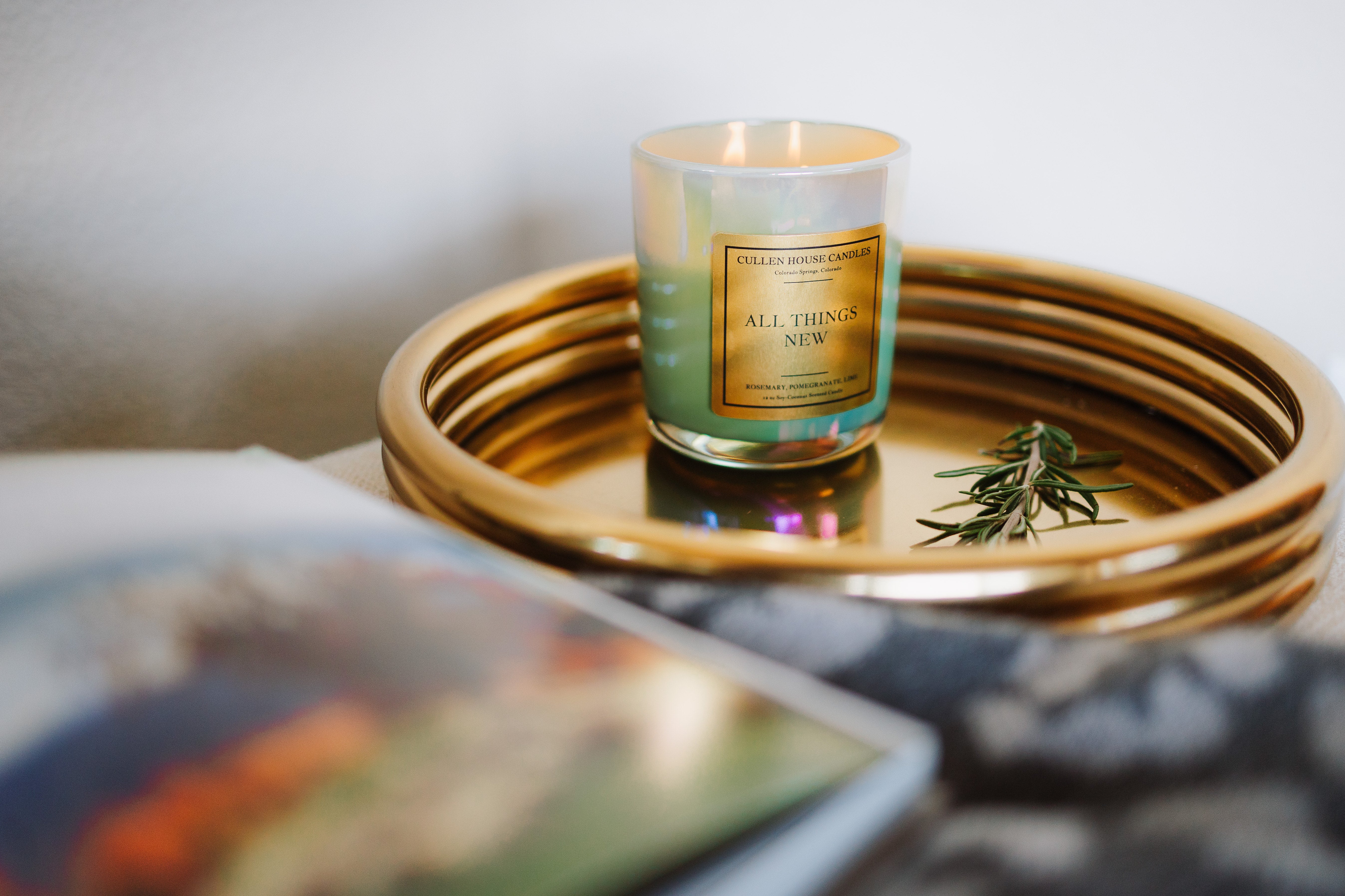 Candle labeled 'All Things New' on a gold tray with a blurred background